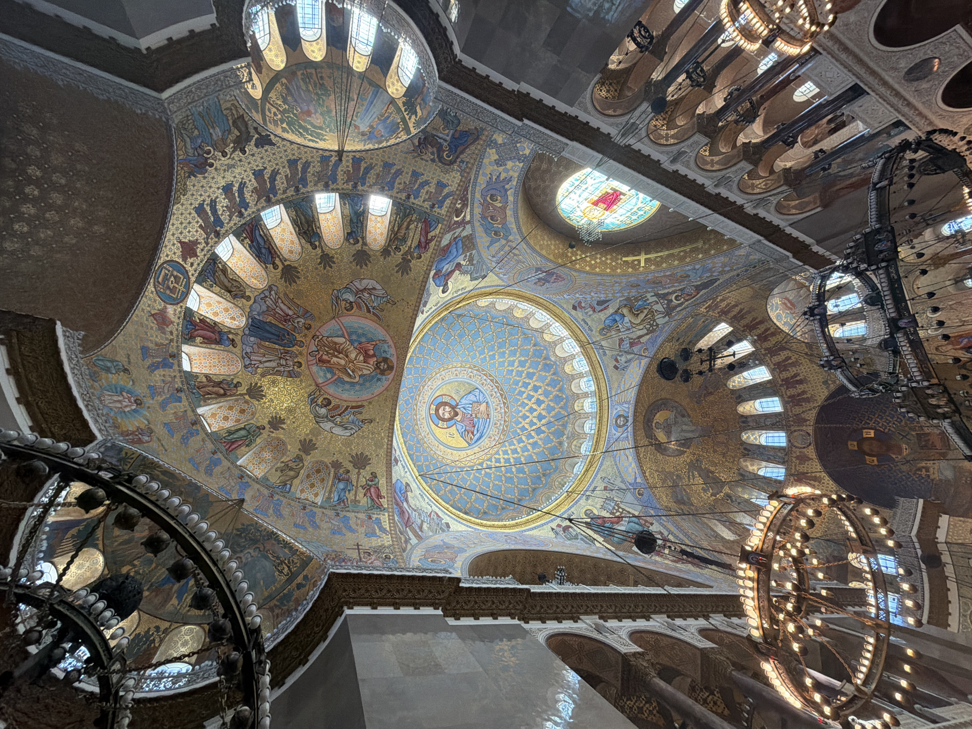The image captures the interior of the Naval Cathedral of Saint Nicholas in Kronstadt, Russia, specifically focusing on the ornate ceiling. The ceiling is adorned with intricate mosaics depicting religious figures and scenes. Multiple domes are visible, each with different patterns and colors. A large chandelier hangs from the ceiling, partially obscuring some of the mosaic work. Architectural details such as arches, columns, and decorative cornices are also visible. The lighting is a mix of natural light coming through windows in the domes and artificial light from the chandeliers.