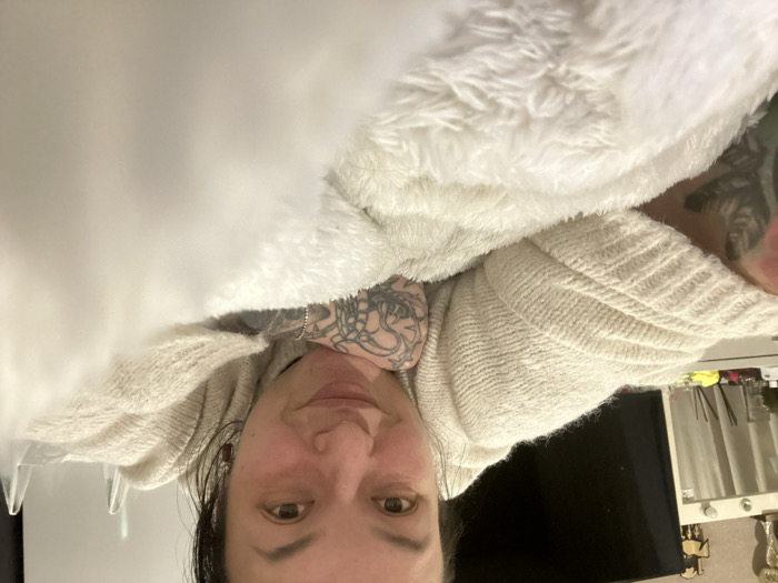 The image displays a female subject, an adult woman, positioned prone, looking directly into the camera with a slight smile. Her face and upper torso are visible. She has dark hair and dark eyes. She is wearing a light-colored, cream or off-white, knitted sweater. Her visible forearms are extensively covered in dark-ink tattoos, with a distinctive spiderweb pattern identifiable on her left elbow area. A silver-colored bracelet is present on her right wrist. She is resting on a thick, white, fluffy blanket which occupies the lower and central foreground. The background reveals an indoor domestic setting in the city of Chaikovsky, Russia. To the left, a white vanity dresser is visible, featuring an illuminated mirror with lights along its top edge. Objects on the dresser include small bottles, a reed diffuser, a small golden trophy or figurine, and a wooden decorative item resembling a horse. Behind the dresser, a large, dark flat-screen television is mounted on a wall.