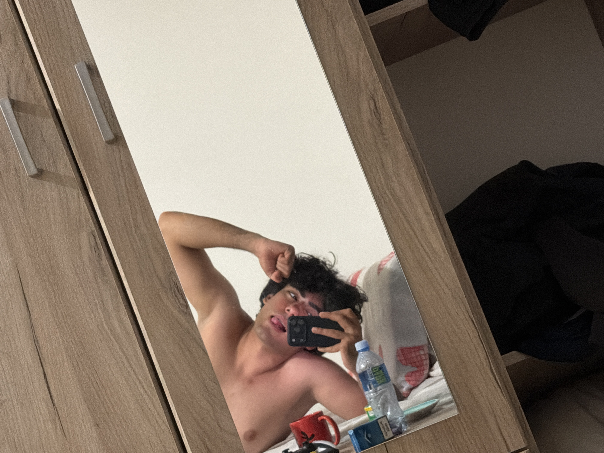 The image depicts a selfie taken in a bedroom. The person taking the photo is a shirtless young man with dark curly hair. He is holding a black smartphone and sticking his tongue out. The reflection shows a bed with a patterned pillow and a nightstand. On the nightstand are a water bottle, a red mug, a pack of cigarettes, and a small object. The mirror is attached to a wooden wardrobe. The location is not explicitly stated in the image, and therefore I cannot confirm if it's in Podgorica, Montenegro.