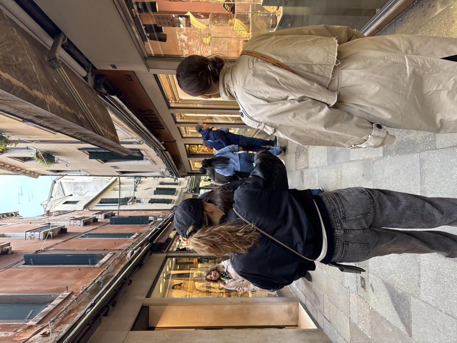 The image displays a narrow pedestrian alleyway, characteristic of a *calle* in Venice, Italy. The ground is paved with light grey stone. Multi-story buildings with aged facades, reddish-brown on the left and cream-colored on the right, line both sides of the street, featuring dark green window shutters on upper floors.
In the immediate foreground, two individuals are walking away from the camera. The person on the left wears a black baseball cap, a black bomber jacket over a white shirt, and faded dark grey jeans. Their light brown hair is styled in a ponytail, and a thin gold chain strap from a small black bag is visible across their back. The person on the right wears a light beige trench coat with a visible brown strap across their back; their hair is pulled back into a bun.
Further down the street, on the left, a woman with brown, shoulder-length hair, wearing a light coat and a patterned scarf, looks towards the foreground from within a shop entrance. On the right, a man in a dark blue jacket leans into a shop window, observing the displayed items. Another individual in a light blue top stands nearby.
Shopfronts with large glass display windows are present on both sides. The shop on the right has an awning labeled "FAURIS" and displays what appear to be jewelry or ornate objects in illuminated cases. The shop on the left shows mannequins with clothing, with the partial word "EVELEEN" visible. Fabric awnings extend over both sets of shop windows. Overhead, utility lines are visible against a light, overcast sky.