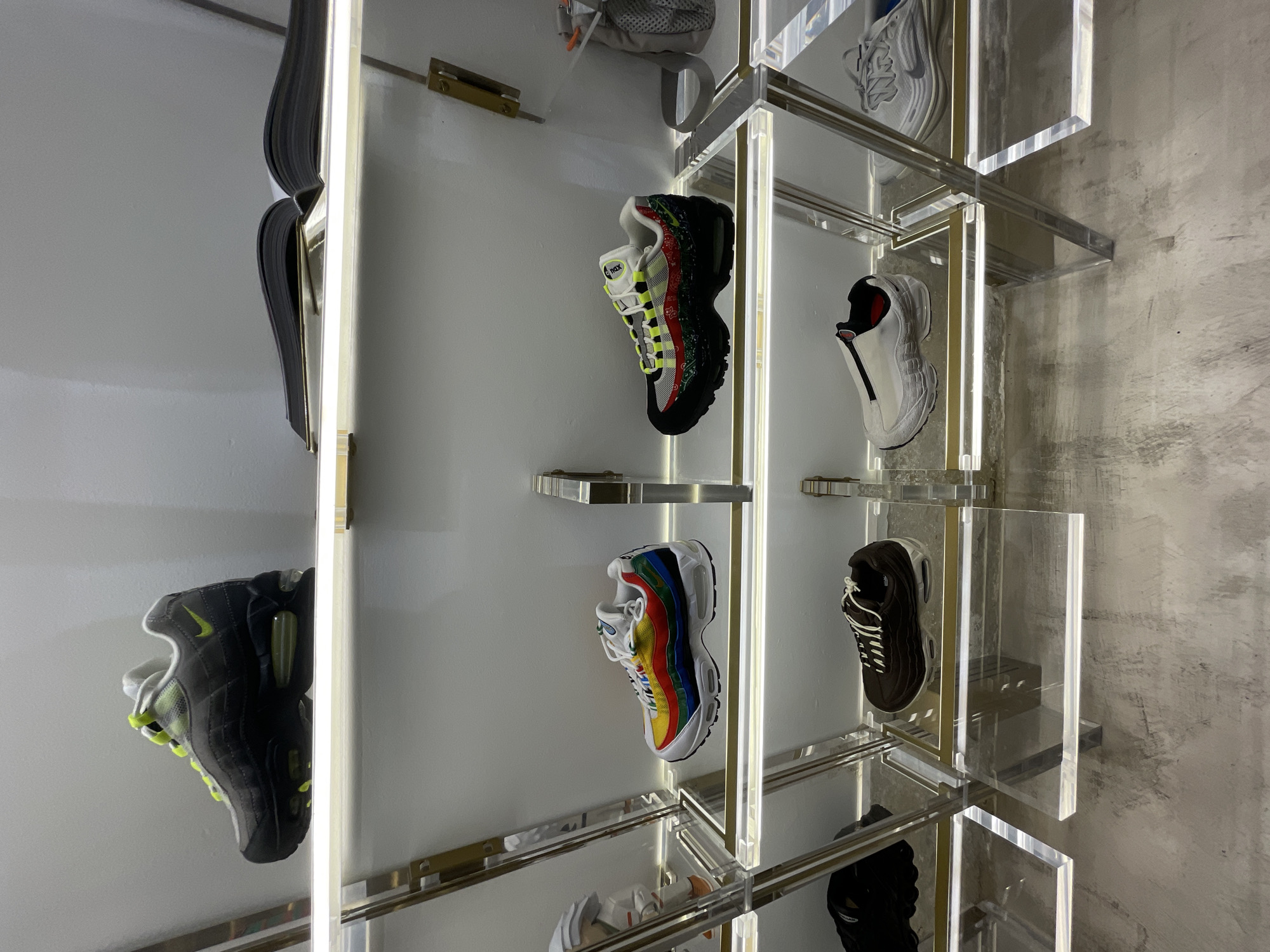 This vertical shot captures a modern retail display showcasing various athletic sneakers, predominantly Nike Air Max 95 models, within what appears to be a store or exhibition space.
The central subjects are multiple pairs of sneakers, each presented on individual clear acrylic or glass shelves. These shelves are supported by thin, gold-toned metal frames, which are vertically illuminated by integrated LED strips, casting bright lines of light.
On the left, a black sneaker with vivid neon yellow-green accents is partially visible at the top and fully shown at the bottom. In the upper middle, a black sneaker features distinctive layered side panels adorned with red, green, yellow, and blue stripes. The tongue of this shoe clearly displays "Air Max" in white text on a green patch. Directly below it, another Nike Air Max 95 boasts full rainbow-colored stripes across its white upper. To the right, a white or light gray sneaker with red accents and a dark brown or black sneaker with white laces are displayed on shelves, with a textured concrete wall visible in the background to their right. Other shoes are partially visible or reflected in the background.
The setting is an indoor, contemporary retail environment, characterized by clean lines, transparent materials, and strong artificial lighting designed to highlight the merchandise. No people are visible, and no active event is taking place; the image captures a static product display. The bright illumination suggests an indoor setting, likely during business hours. While the location context is provided as Barcelona, Spain, there are no specific visual cues within the image itself to identify this city. The display exudes a sleek, modern, and curated aesthetic, emphasizing the design and variety of the footwear.
