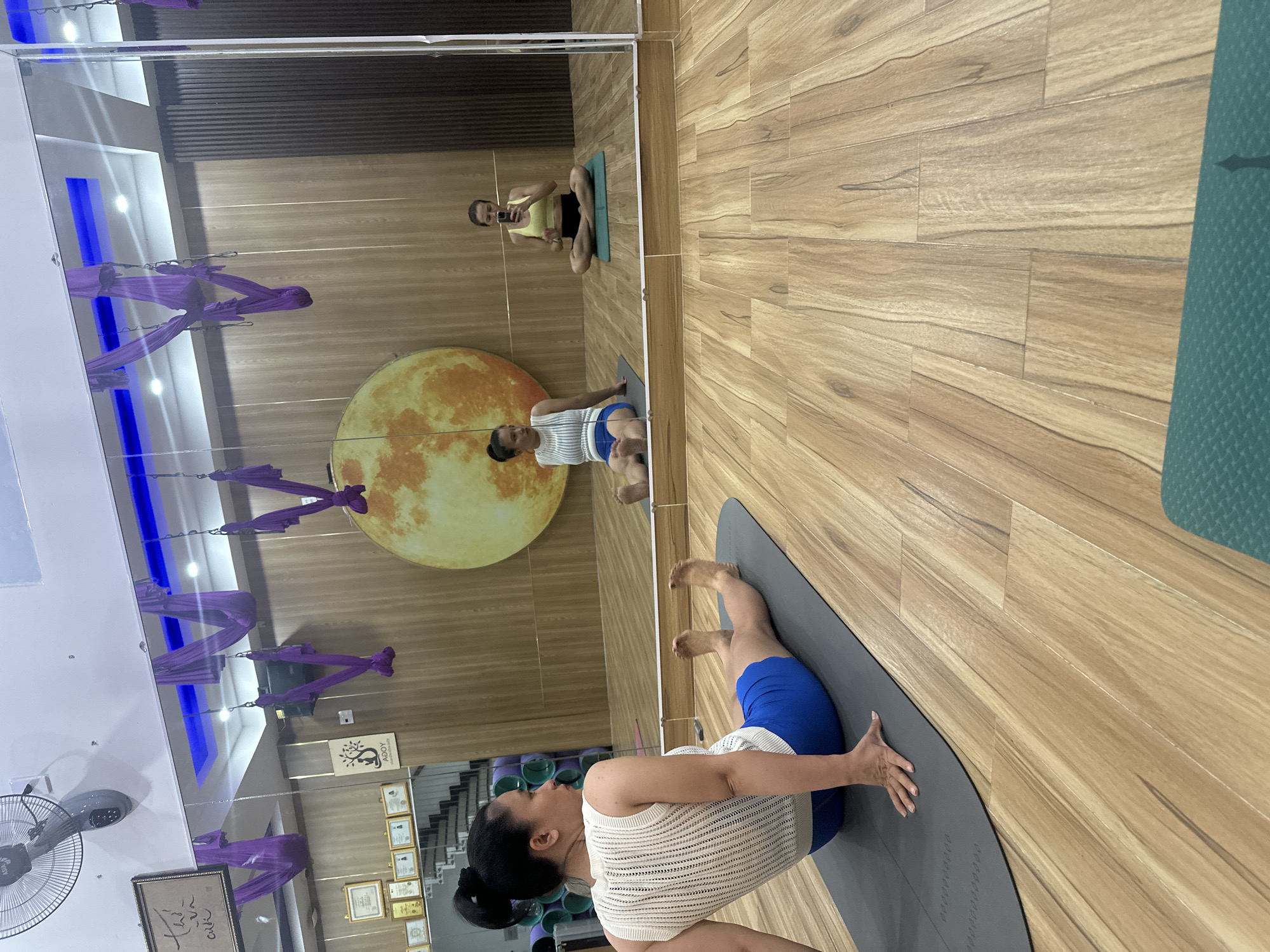 A woman is performing a yoga pose in a studio in Bac Nha Trang Ward, Vietnam. She is on a grey yoga mat, wearing a white top and blue shorts. Behind her is a mirror reflecting the studio, which includes aerial yoga silks hanging from the ceiling, a large moon image on the wall, and a view of another person sitting on a mat, looking at a phone. The room is well-lit, with a wooden floor. There are framed certificates on the wall.