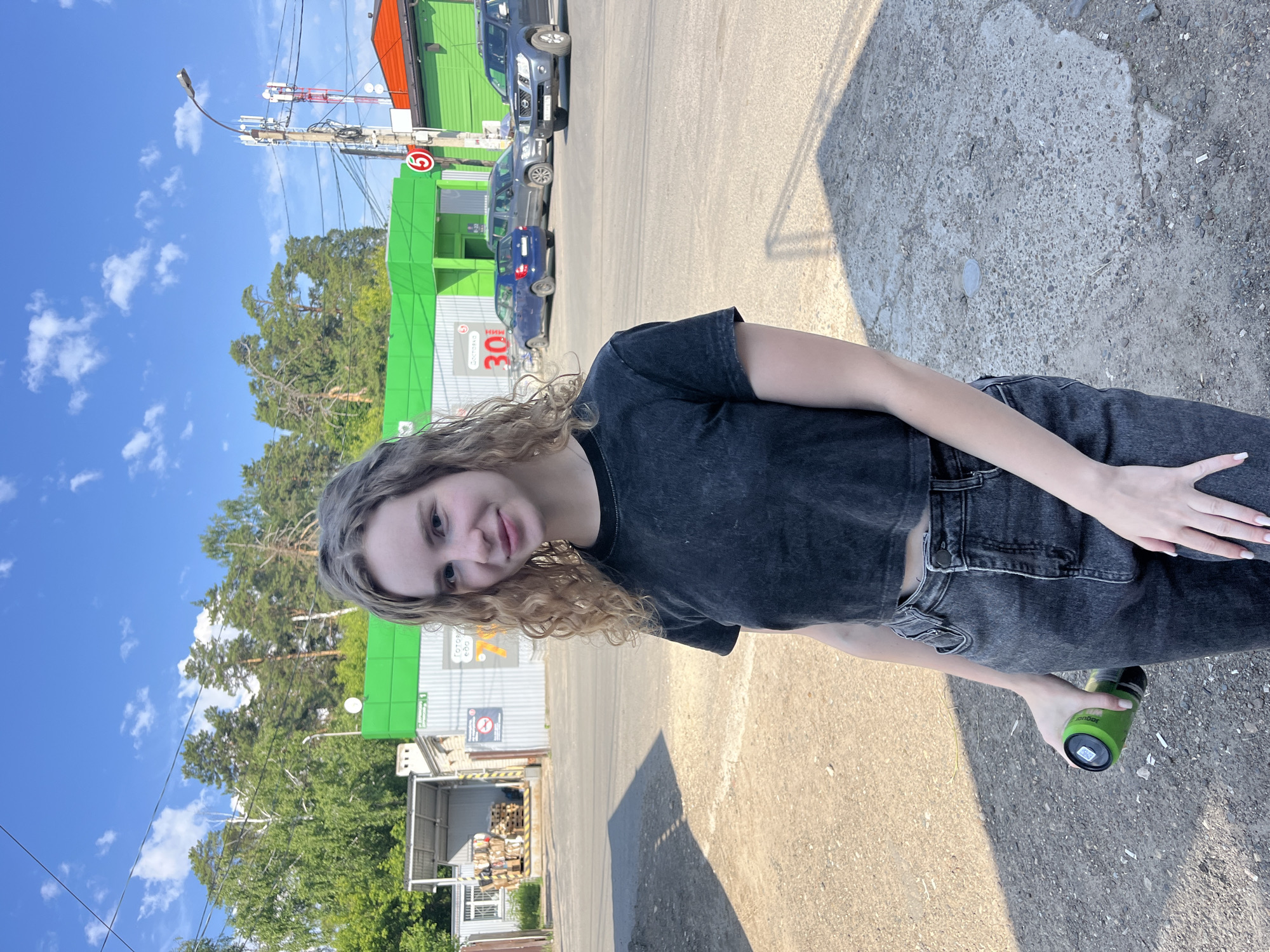 A young woman with curly blonde hair stands in front of a row of buildings under a bright blue sky with scattered clouds. She is wearing a dark gray t-shirt and dark gray jeans, and holding a green can in her right hand. The buildings appear to be shops or businesses, with one notable for its bright green facade and another with a red roof. Several cars are parked in front of the buildings. In the background, tall green trees are visible, and further back, a utility pole with multiple antennas is present. The ground in the foreground is a mix of gravel and concrete, with some shadows cast by the sunlight. This scene appears to be set in Vasilyevo, Russia, possibly in a commercial or industrial area.