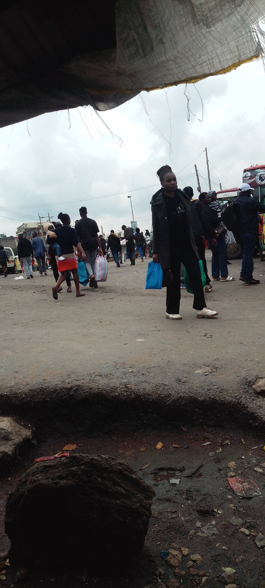 The image depicts a busy, overcast daytime scene in an urban or semi-urban area, likely a market or transport hub in Nairobi, Kenya. The foreground features rough, unpaved ground with a large dark rock on the left and scattered debris including small stones and muddy patches, suggesting recent rain. A dark, frayed overhead tarp or canopy partially covers the top of the frame.
Numerous people are visible across the midground and background, indicating significant pedestrian traffic. A woman stands prominently towards the center-right, facing the viewer with a neutral expression. She is dressed in a dark coat, dark trousers, and white sneakers, holding two bright blue plastic bags. Her hair is styled in dark braids or locs pulled upwards. Behind her and to her left, other individuals are walking, some carrying bags of various sizes and colors, including a large striped bag and more blue plastic bags. One person in the mid-left appears to be carrying a child.
In the background, more pedestrians are visible, along with parts of buildings and utility poles with power lines. A red and white bus, characteristic of Kenyan "matatus," is partially seen on the far right, parked or moving slowly. The sky is entirely overcast, suggesting a cloudy day without direct sunlight. People generally appear focused on their movement, without strong emotional displays. No legible text is discernible on any signs, vehicles, or clothing. The scene conveys a sense of everyday activity and bustling urban life.