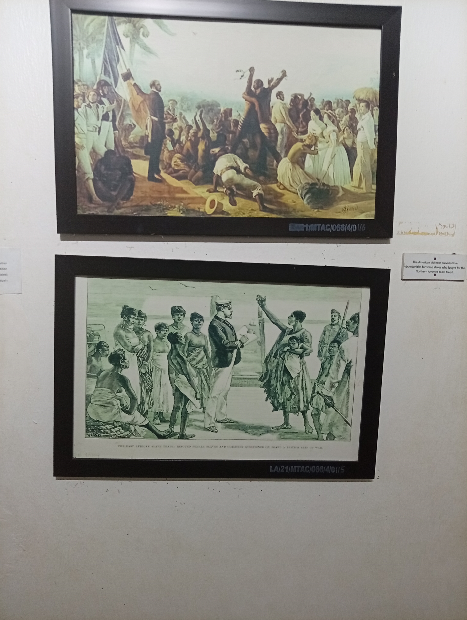 The image shows a display of two framed prints on a wall, likely within a gallery or museum setting in Unknown, Unknown.
The top print is a color painting depicting a historical scene with a crowd of people. Some individuals appear to be enslaved, while others are dressed in formal attire, suggesting a historical context related to emancipation or abolition.
The bottom print is a monochrome illustration, possibly a lithograph, titled "THE EAST AFRICAN SLAVE TRADE. RESCUED FEMALE SLAVES AND CHILDREN QUESTIONED ON BOARD A BRITISH SHIP OF WAR." It portrays a group of women and children, presumed to be rescued slaves, being questioned by men in uniform, presumably British officers.
Textual information is also present, including reference codes "LA/21/MTAC/066/4/0115" and "1/MTAC/066/4/01/6" below the respective prints, and a placard stating, "The American civil war provided the opportunities for some slaves who fought for the Northern America to be freed." The environment appears to be a well-lit, indoor space, with the wall painted in a light color.