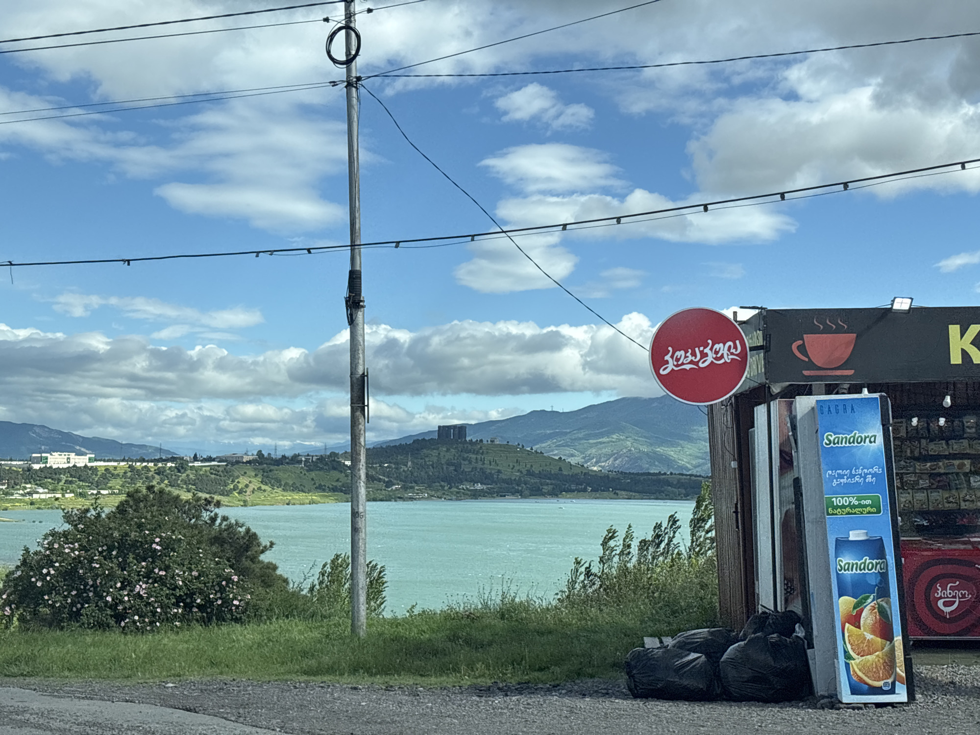 The image depicts a roadside scene near Tbilisi, Georgia, overlooking a lake. In the foreground, there's a small commercial structure with a red circular sign in Georgian script and a sign advertising "Sandora" juice. Black trash bags are piled in front of the structure. An electric pole runs vertically, cutting through the scene. In the background, a large body of water stretches towards rolling green hills and mountains, capped with scattered clouds in a blue sky. A prominent building sits atop one of the hills. Vegetation is visible along the roadside.