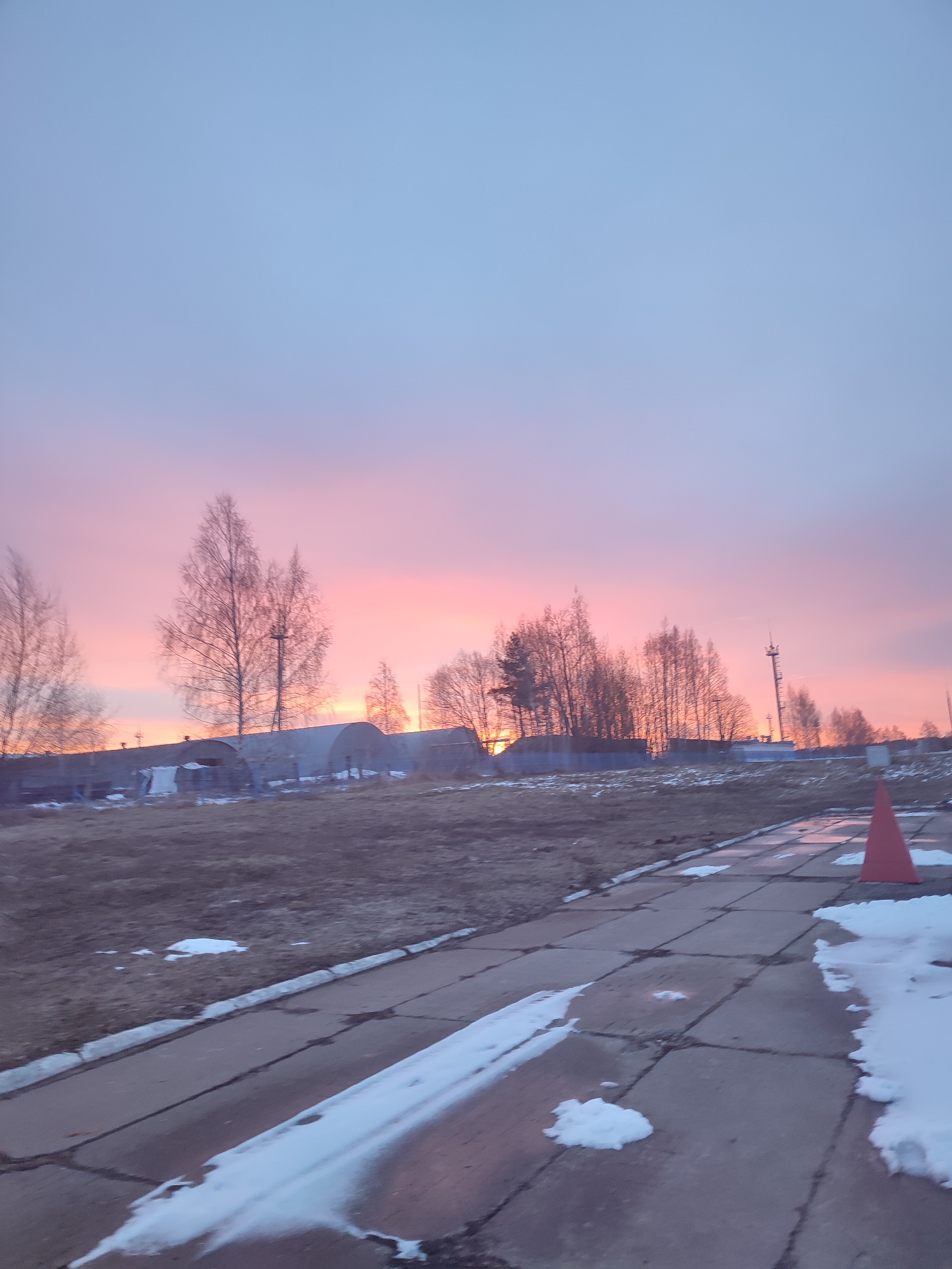 The image depicts an outdoor scene during a colorful sunset. The sky transitions from light blue to pink and orange hues. Silhouetted trees and buildings are visible against the bright horizon. In the foreground, there's a paved walkway with patches of snow and an orange traffic cone on the right. The ground is a mix of grass and dry vegetation. The location appears to be on the outskirts of a town or rural area, possibly in Unknown, Unknown, but no clear markers exist in the image.