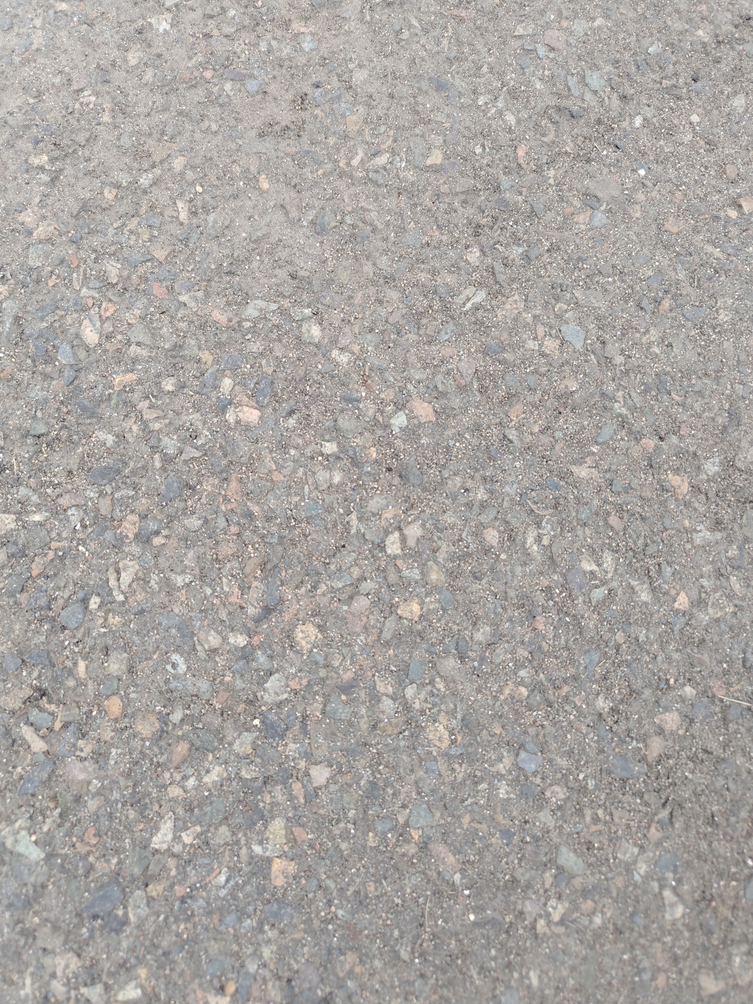 The image shows a close-up, overhead view of asphalt pavement. The surface is composed of small grey, black, and reddish-brown aggregate mixed with a dark binder. Some larger, irregularly shaped stones are visible within the asphalt. There are no people, structures, or distinct objects present in the frame. The lighting suggests an outdoor setting, likely during daylight hours. The image's context places it in the city of Stepnoe, Russia, though no specific landmarks or environmental features are identifiable within the image itself to confirm this location.