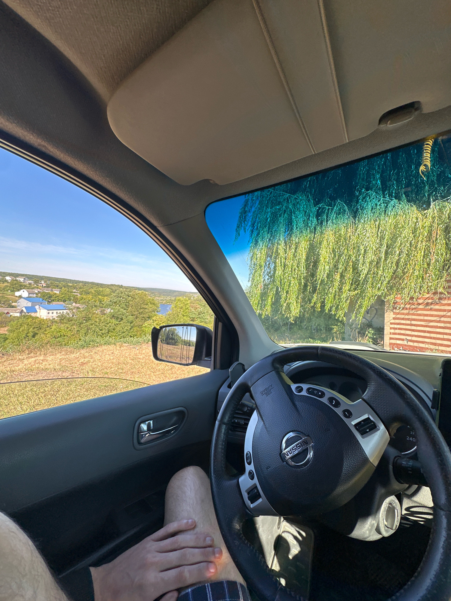 The image depicts the interior of a car, viewed from the perspective of the driver. A person's legs and hand are visible in the foreground, indicating they are in the driver's seat. The steering wheel, with a Nissan logo, is centrally located. Through the open driver's side window, a rural landscape is visible, featuring rolling hills covered in green vegetation and a cluster of houses with blue roofs. A small body of water can be seen in the distance. A side mirror reflects a similar rural scene. The windshield shows a large weeping willow tree casting shadows, and a brick structure. The sky is clear and blue. The image appears to be taken in Makiv, Ukraine, given the context provided, likely on a sunny day.
