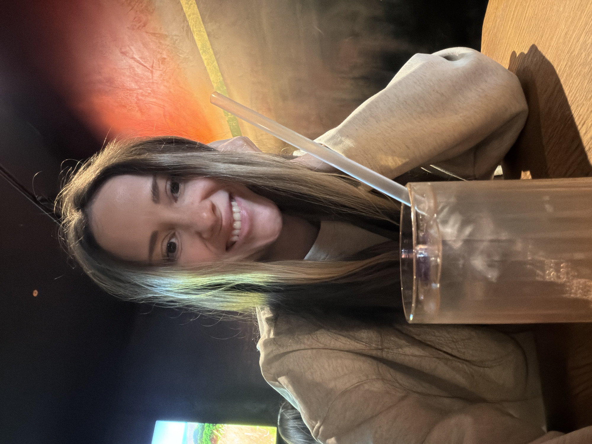 A female subject, appearing to be in her twenties to thirties, is seated indoors, smiling directly at the camera. She has long, light brown hair with lighter streaks. She is wearing a light beige or grey long-sleeved garment, with her left elbow resting on a wooden table and her left hand supporting her head. In the foreground, a tall, clear glass filled with clear liquid and ice cubes, containing a white or translucent straw, sits on the wooden table. The background is dimly lit; a bright screen displaying a colorful, stylized landscape with green hills and a blue sky is visible to the left. To the right, a textured wall exhibits an orange-red gradient with a diagonal yellow stripe. The specific location within Khimki, Russia, cannot be identified from the provided image.