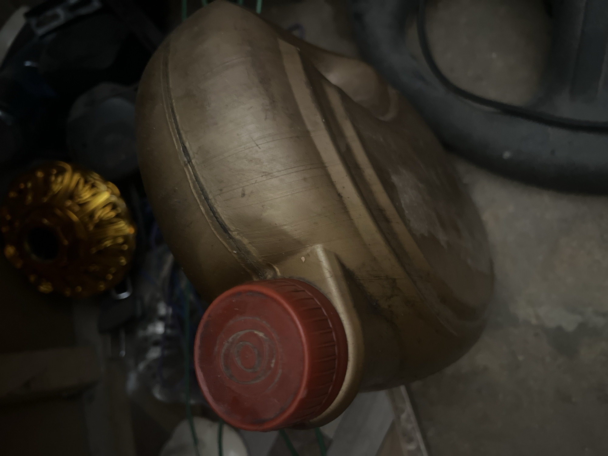 The image shows a close-up of a large, tan plastic jerrycan with a red cap. The jerrycan is resting on a textured surface, possibly concrete or tile, in what appears to be an indoor storage area. In the background, various items are visible, including what looks like a golden decorative object with a circular pattern, a dark spherical object, and other indistinct objects and materials. The overall lighting is dim, and the scene is somewhat cluttered. The location is identified as Jalingo, Nigeria.