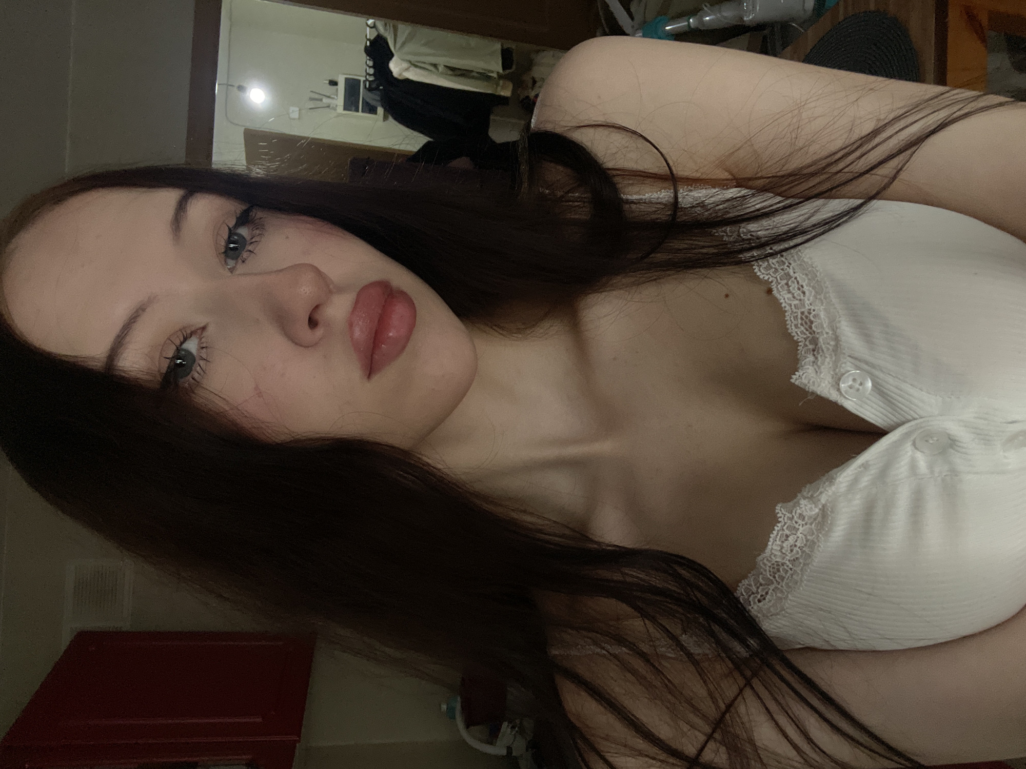 The image is a close-up, waist-up selfie of a young woman with long, dark hair. She is wearing a white ribbed tank top with lace trim and two buttons down the front. Her makeup includes eyeliner and lip gloss. The background appears to be an indoor residential setting. To the left, a portion of a red cabinet is visible. The right side of the background shows a doorway with clothes hanging on a rack and a shelf unit. A light fixture is visible in the upper right portion of the background. The image's lighting is dim and appears to be from an overhead source. The woman's gaze is directed towards the camera. The location is Votkinsk, Russia.