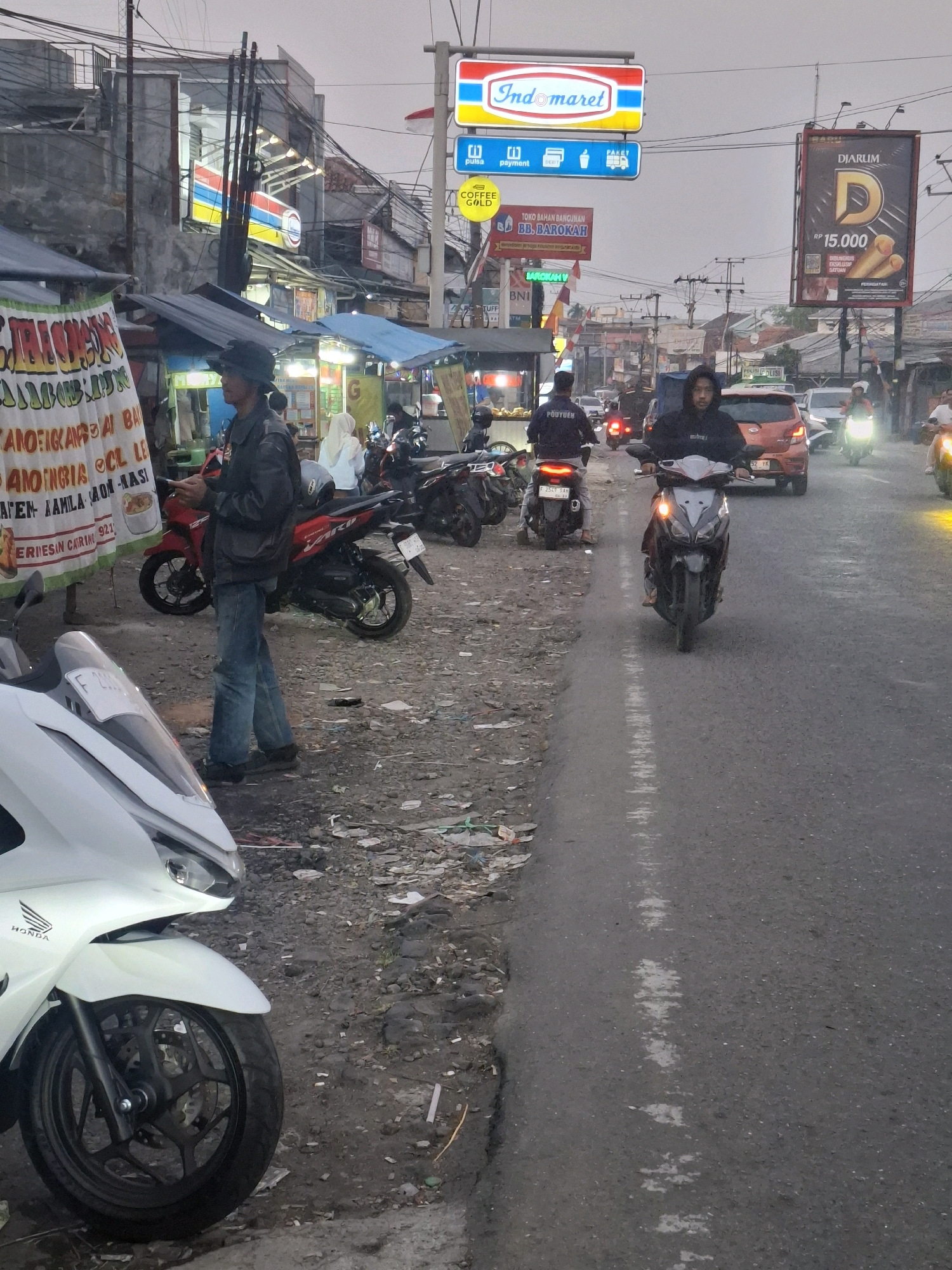 A street scene in Cikaroya, Indonesia, shows a moderately busy road with several motorcycles and a few cars. The sky is overcast, suggesting it may be early morning or late afternoon. On the left side of the road, there are several street vendors with their stalls set up under awnings. One stall has a large banner with Indonesian text. A man wearing a black jacket and jeans stands near the stalls, looking at his phone. Several motorcycles are parked or being ridden on the road.
In the background, there are buildings, including a prominent Indomaret convenience store with its distinctive red and blue signage. Another large billboard for Djarum cigarettes is also visible. The road itself has a white line marking its center. The ground on the side of the road is unpaved and appears to be littered with debris. One person is riding a motorcycle in the foreground, and another is in the middle ground. The overall impression is a typical urban street scene in Indonesia with commercial activity and traffic.