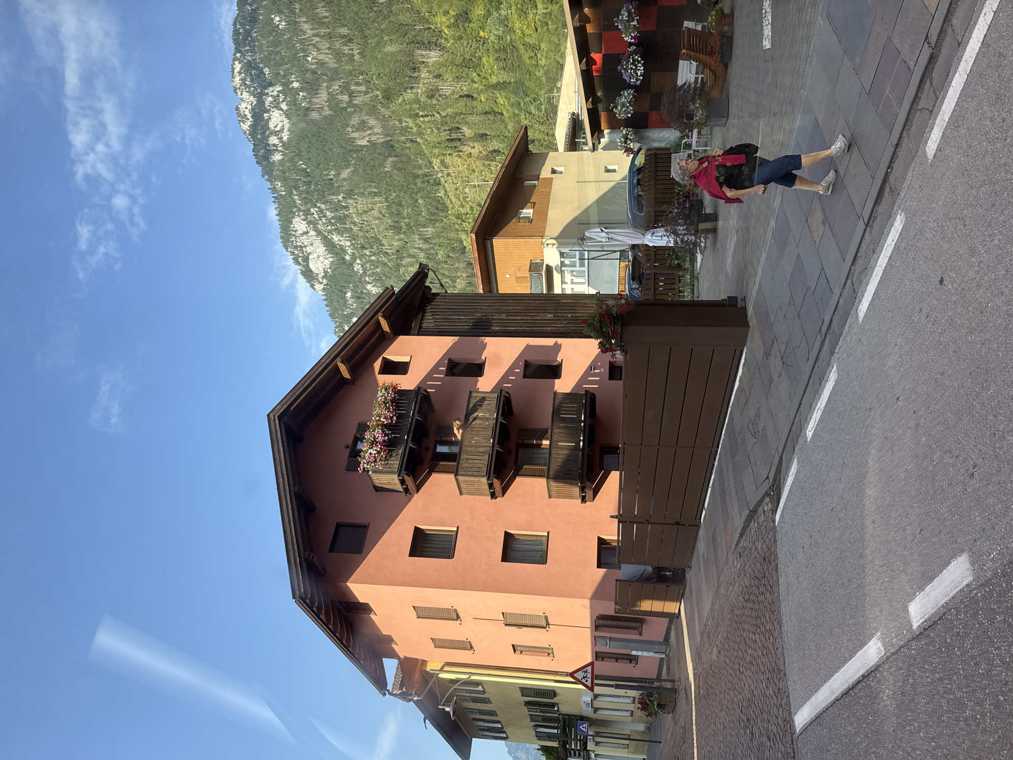The image shows a street scene in Predazzo, Italy. A woman with gray hair wearing a red scarf over a black shirt and blue capri pants walks on a sidewalk. She is carrying a black bag. The street is paved with asphalt and has white lane markings. A cobblestone sidewalk runs parallel to the street, and beyond that, a paved walkway with a wooden fence. Buildings line the street, including a salmon-colored building with balconies adorned with flowers and a yellow building with shutters. In the background, a steep, forested mountain rises under a clear blue sky with some wispy clouds. A traffic sign depicting children crossing is visible near the yellow building. The overall scene suggests a quiet day in a town situated in a mountainous region.