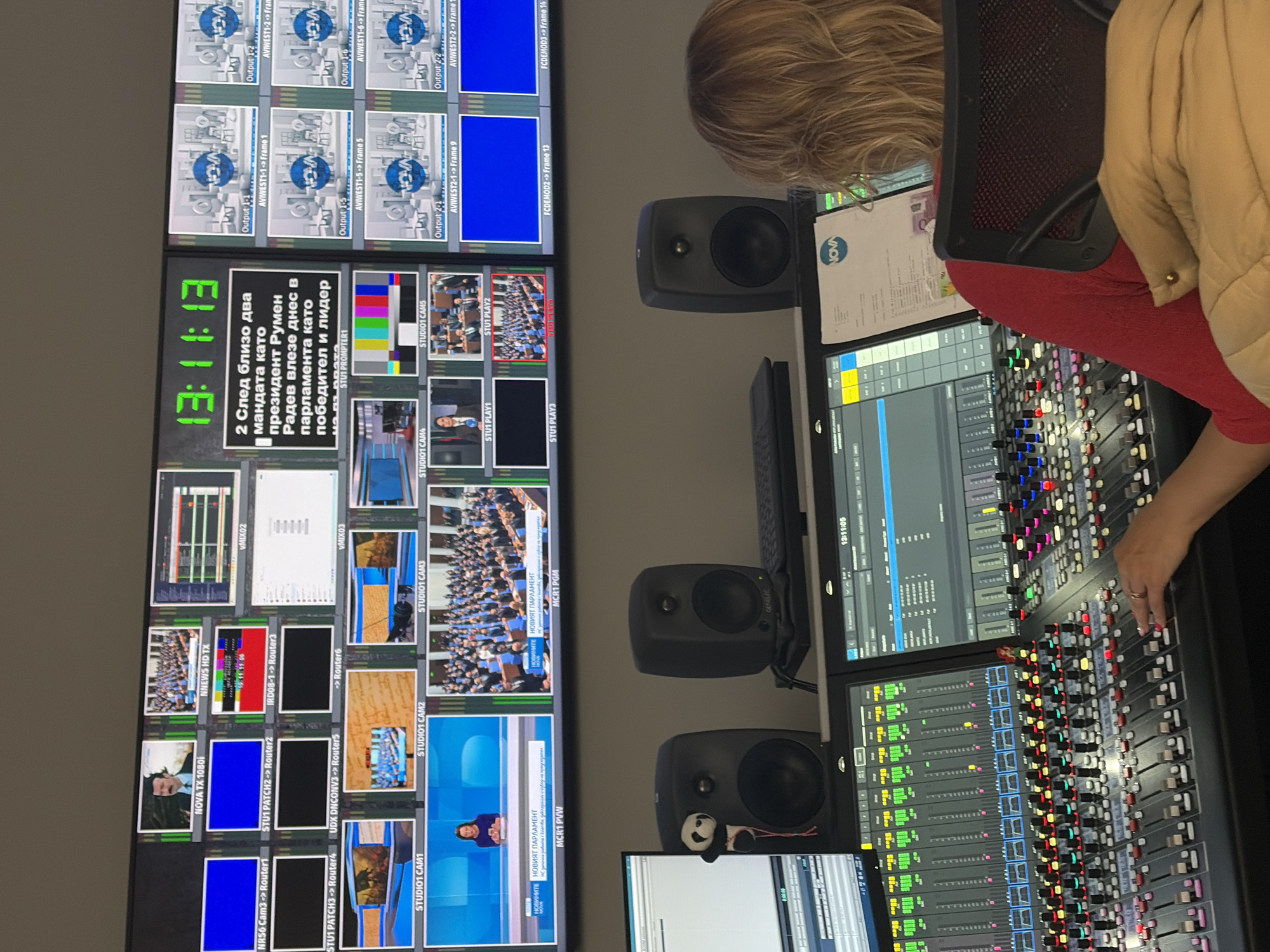 The image depicts an operator working in a television broadcast control room, likely located in Busmantsi, Bulgaria. A person with blonde hair is seen from behind, seated in a black chair with a red backrest, operating a large audio mixing console with numerous colorful faders and buttons. Their right hand is actively engaging with the controls.
To the left of the operator, a massive multi-monitor display wall showcases a complex array of video feeds, graphics, and technical information. One prominent screen shows a digital clock displaying "13:11:13" and a Bulgarian news headline that translates to "2 Following nearly two terms as president Rumen Radev entered parliament today as winner and leader." Other monitors display various video sources, including several labeled "STUDIO1 CAM1" through "STUDIO1 CAM5," showing views of what appears to be a parliamentary session or a large assembly. Further technical labels such as "NOVA TX 1080i", "NNEWS HD TX", "AVIWEST1-1 -> Frame 1", "MCR1 PVW", and "MCR1 PGM" are visible, indicating video routing and program monitoring. Some screens show color bars or blue placeholder screens.
On the desk, alongside the mixing console, are two Genelec brand studio monitor speakers, with a small panda plush toy adorning the lower one. Computer screens display audio waveform software interfaces, and one shows a digital clock at "13:11:05". A document with the "NOVA" logo is also visible on the desk. The scene captures the active, technical environment of a live broadcast or news production.