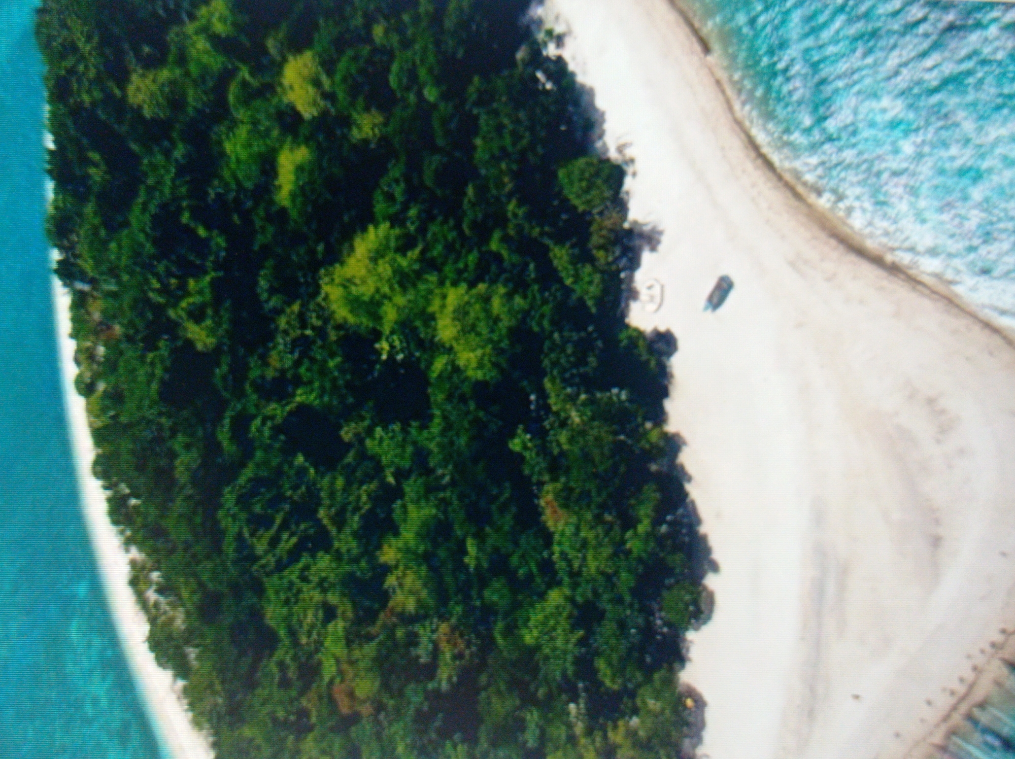 The image displays an aerial view of a tropical island. The island is characterized by a lush, green forested interior and a white sandy beach that curves along the coastline. The ocean surrounding the island is a vibrant turquoise, with visible waves breaking on the shore. On the beach, there is a small, dark-colored boat pulled up onto the sand, and what appears to be a white lounge chair or small structure. The image captures a tranquil and remote natural setting. The image does not show or depict the city of Mukachevo, Ukraine.