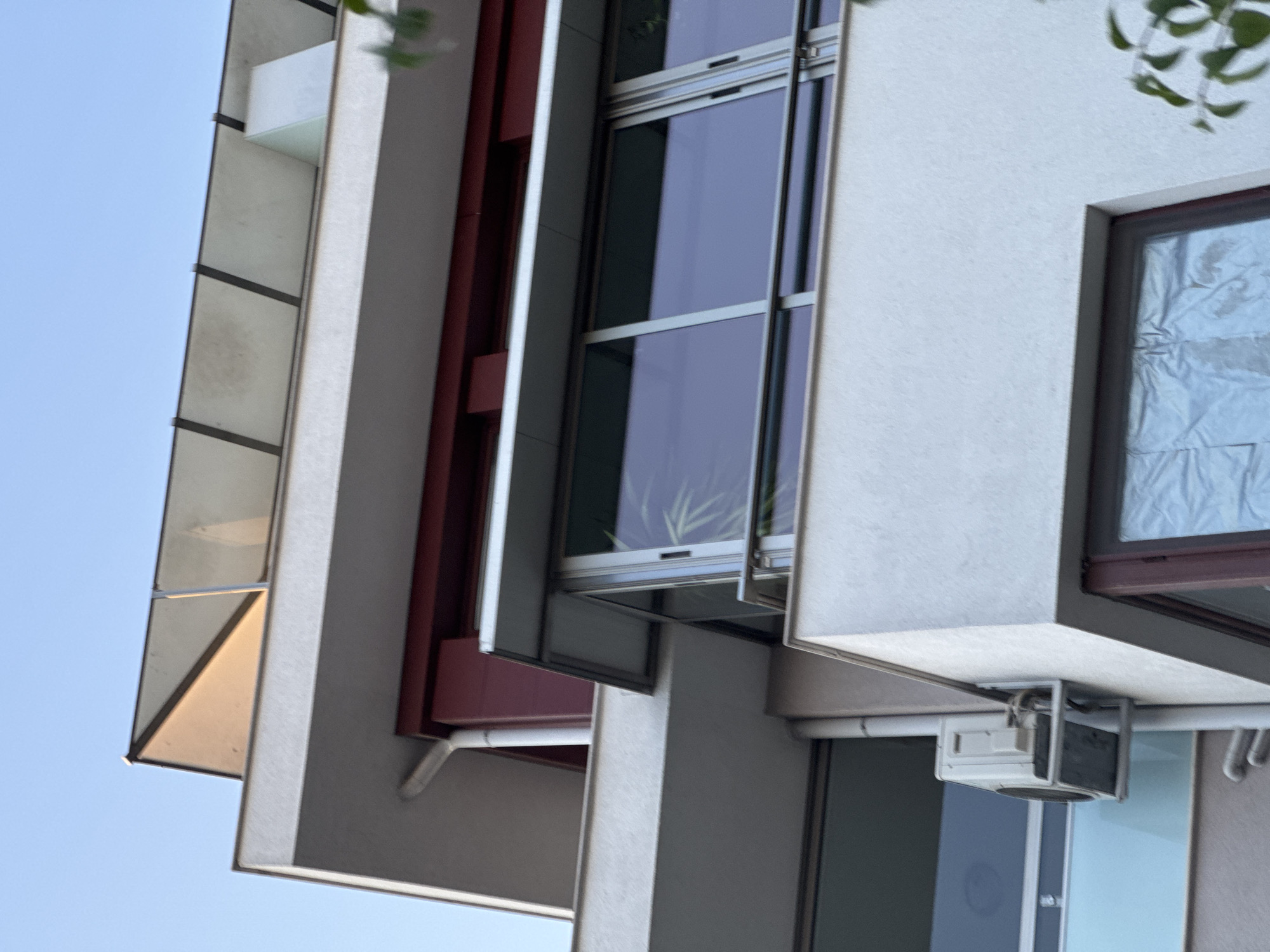 The image shows a low-angle exterior view of a modern building, possibly an apartment complex, under a clear blue sky. The building features multiple levels with contrasting architectural elements. The top portion of the building has a slanted roof structure with what appear to be solar panels or skylights. Below this, there's a dark red horizontal band running across the facade, followed by a section with large windows, a balcony with a metal railing, and an outdoor air conditioning unit mounted on the wall near a white downspout. The building's exterior walls are primarily a light, textured color. The overall impression is one of contemporary residential architecture.