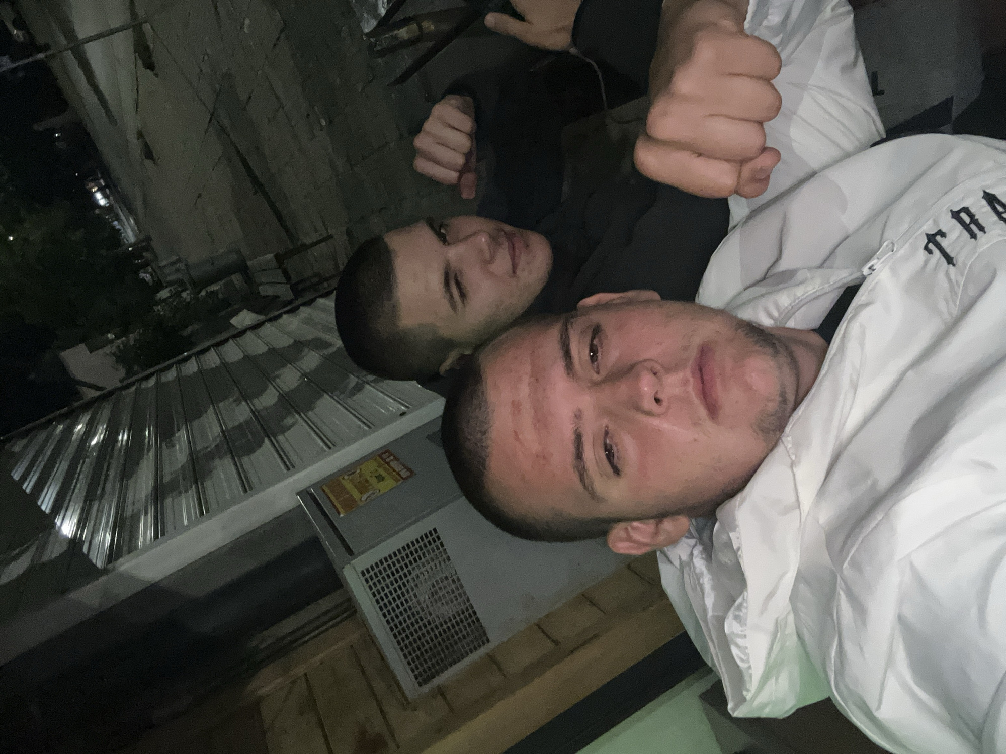 A nighttime, low-angle selfie captures two young adult males in an urban setting within the city of Unknown, Unknown.
The subject in the foreground, positioned left of center, wears a white, slightly reflective, puffer-style jacket with a visible "TRAP" logo in black lettering on the chest. He has a buzz cut and a neutral, direct gaze towards the camera, with visible blemishes on his forehead and light facial stubble. His right hand is loosely clenched and rests on the front of his jacket.
Behind and to his right, the second subject also features a buzz cut and wears a dark-colored (likely black) hooded garment. He winks with his left eye and exhibits a slight smile while holding his right hand in a clenched fist near his face. His left hand is partially visible, appearing to gesture a thumbs-up.
The immediate background on the left reveals vertical wooden paneling. To the right of that, a gray industrial-looking unit with a grated vent is visible, featuring unreadable red and yellow warning labels. Further right, a reflective, corrugated metal roll-down security shutter of a storefront is present. The distant background, visible above and to the right of the subjects, shows a dark, dimly lit urban street with visible asphalt or pavement, dark trees, and faint streetlights in the far distance, indicating nighttime conditions. The overall scene suggests a casual nocturnal encounter or photo opportunity in a quiet urban area.