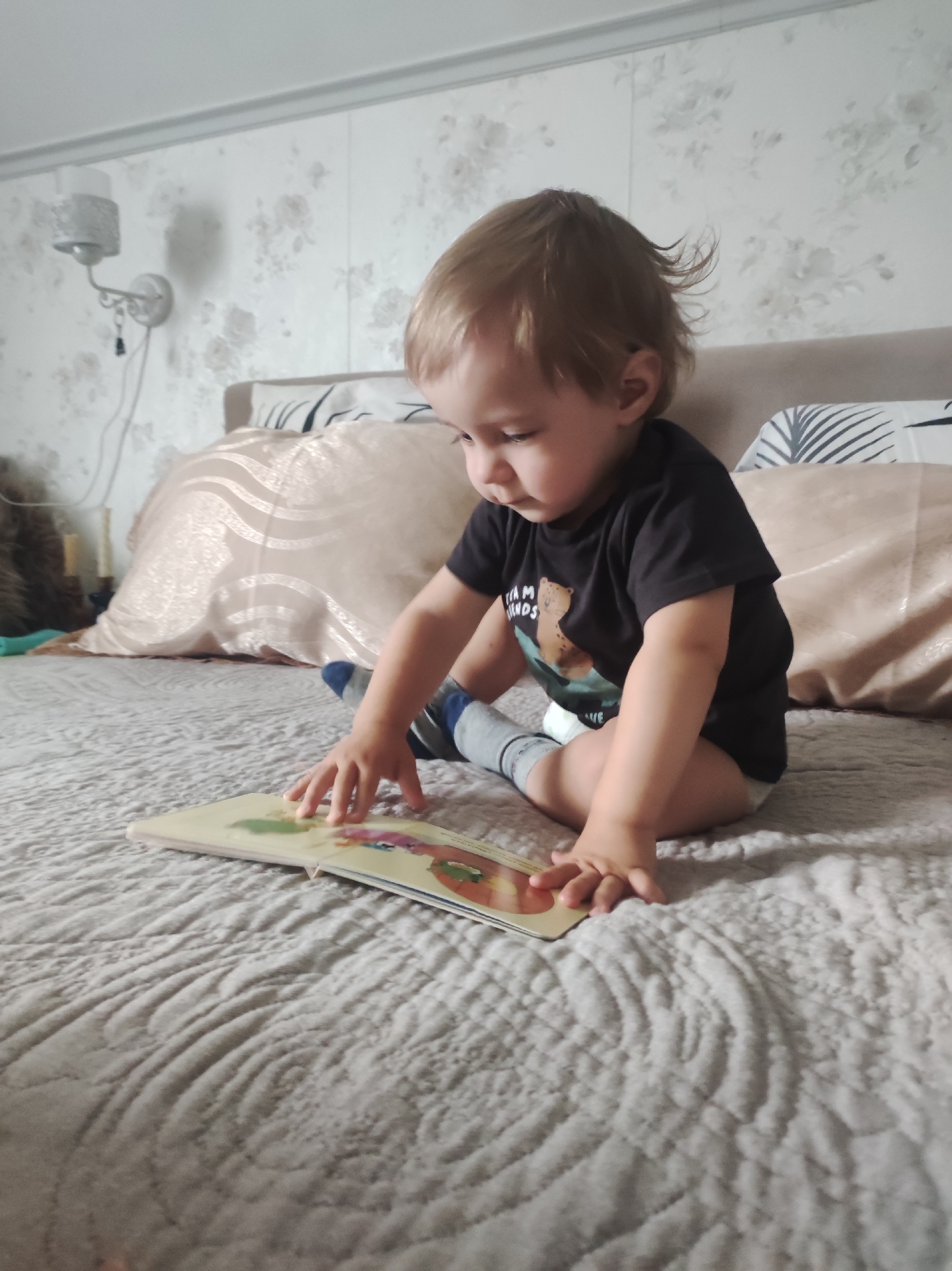 A young child, likely a toddler, with light brown hair, sits attentively on a bed, engrossed in an open children's book. The child wears a black t-shirt with partially visible text "I AM FRIENDS" and "LOVE" alongside a bear illustration, a diaper, and blue and grey striped socks. Their left hand is resting on the colorful illustrations of the book.
The setting appears to be a bedroom. The bed is covered with a light grey quilted bedspread, and several pillows are visible against a beige upholstered headboard. One pillow has a beige case with a wavy pattern, and another has a white case with dark leaf motifs. The wall behind the bed features light-colored wallpaper with a subtle floral design. A white wall-mounted light fixture (sconce) with a decorative shade is visible on the left side of the frame. To the far left, a glimpse of a furry brown object and a turquoise fabric suggests other items on the bed or nearby. The time of day is not evident, but the indoor lighting creates a calm atmosphere for the child's focused activity.