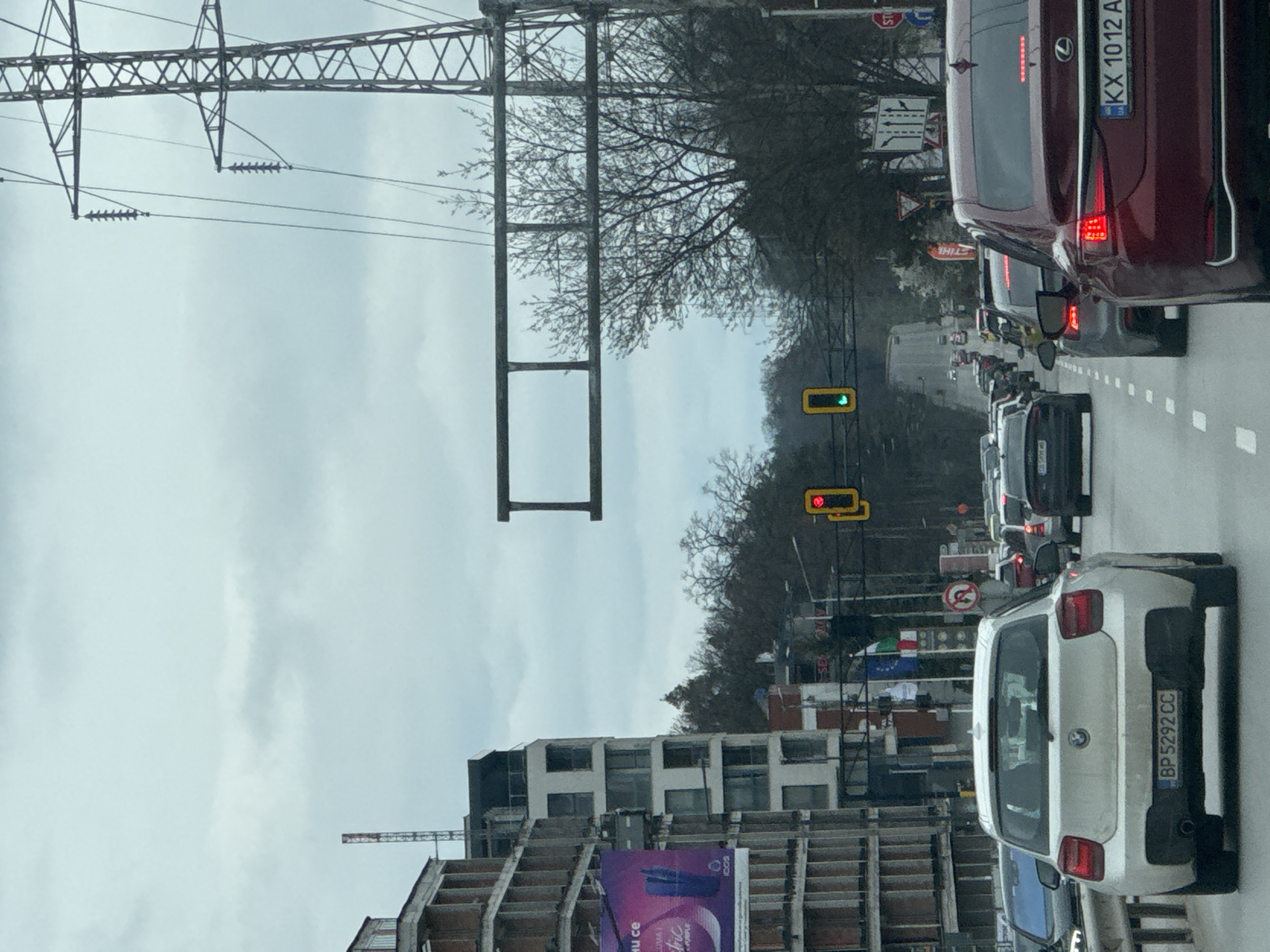 The image depicts a street scene, likely in Sofia, Bulgaria, given the presence of Bulgarian license plates on the vehicles. There is heavy traffic on a multi-lane road. Several cars are visible, including a white hatchback with license plate "BP 5292 CC" and a burgundy Lexus with license plate "KX 1012 AT." Overhead traffic signals are visible, one displaying a green light and another a red light. Construction appears to be taking place on the left side of the frame, with unfinished buildings and scaffolding. Power lines and support structures are also visible overhead. The sky is overcast, and there are trees in the background. No pedestrians are immediately visible.