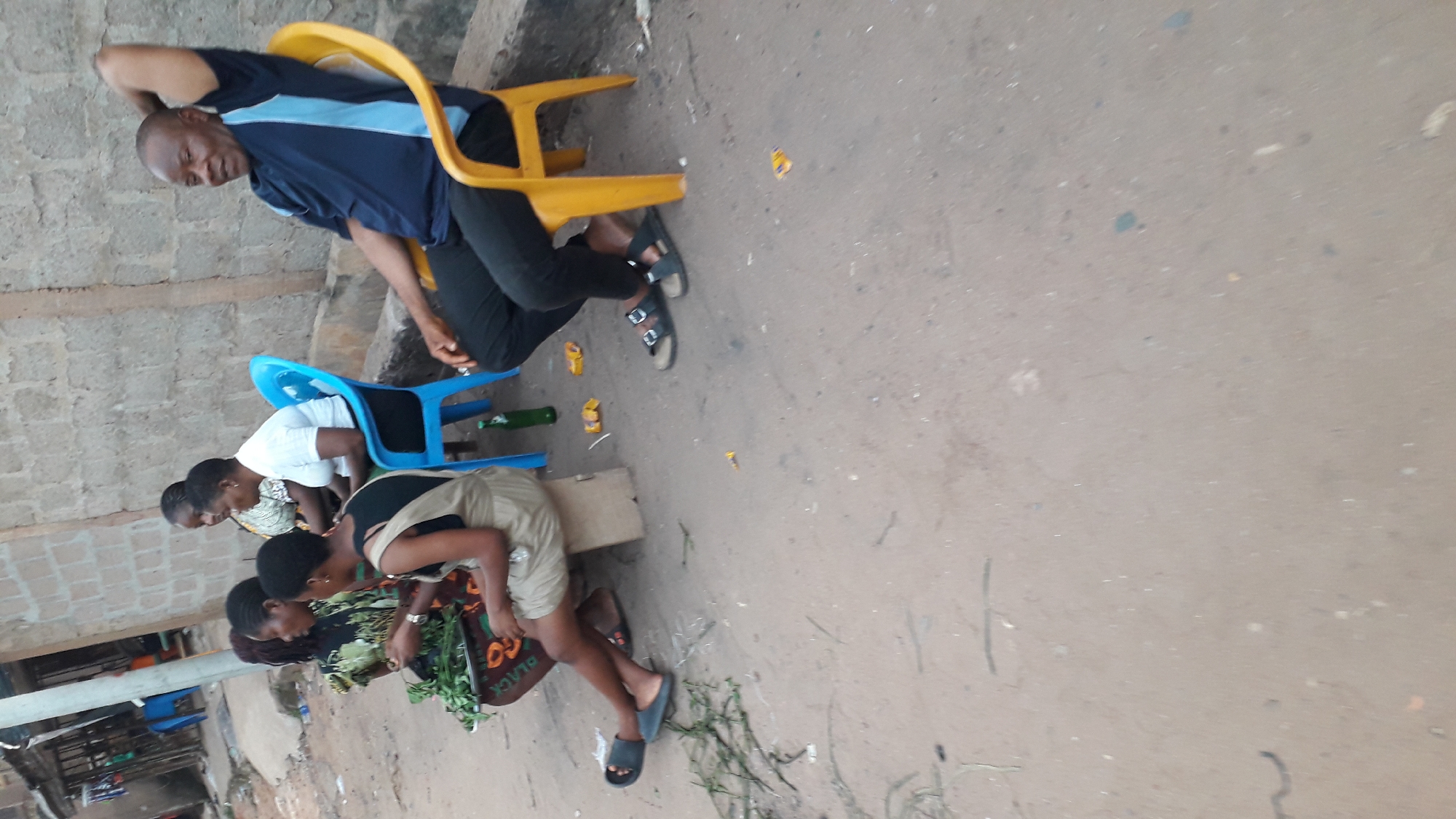 The image shows a street scene in Nkpor, Nigeria. A man is sitting on a yellow plastic chair, wearing a blue and black shirt and black pants. Three women are seated on blue plastic chairs and on a carton box. A woman is bending over a dark basket that holds green vegetables. The ground is paved and there are some small pieces of trash on it. There is a brick wall behind the man.