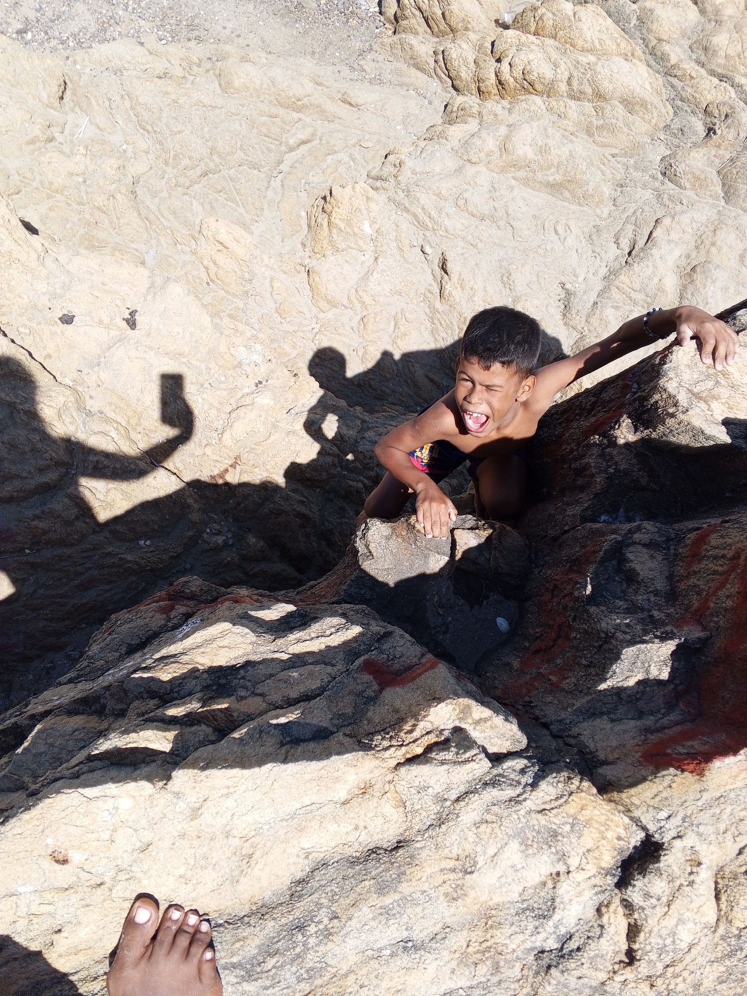 A child is seen climbing on rocks in Juan Griego, Venezuela. The boy is tan-skinned and wearing swim trunks. He is positioned on the rocks with his mouth open in a shout, with his hand gripping a rock. A shadow of a person holding a phone to take a picture is visible on the rocks. A foot is visible in the bottom left corner of the image. The rocks are tan and off-white in color.