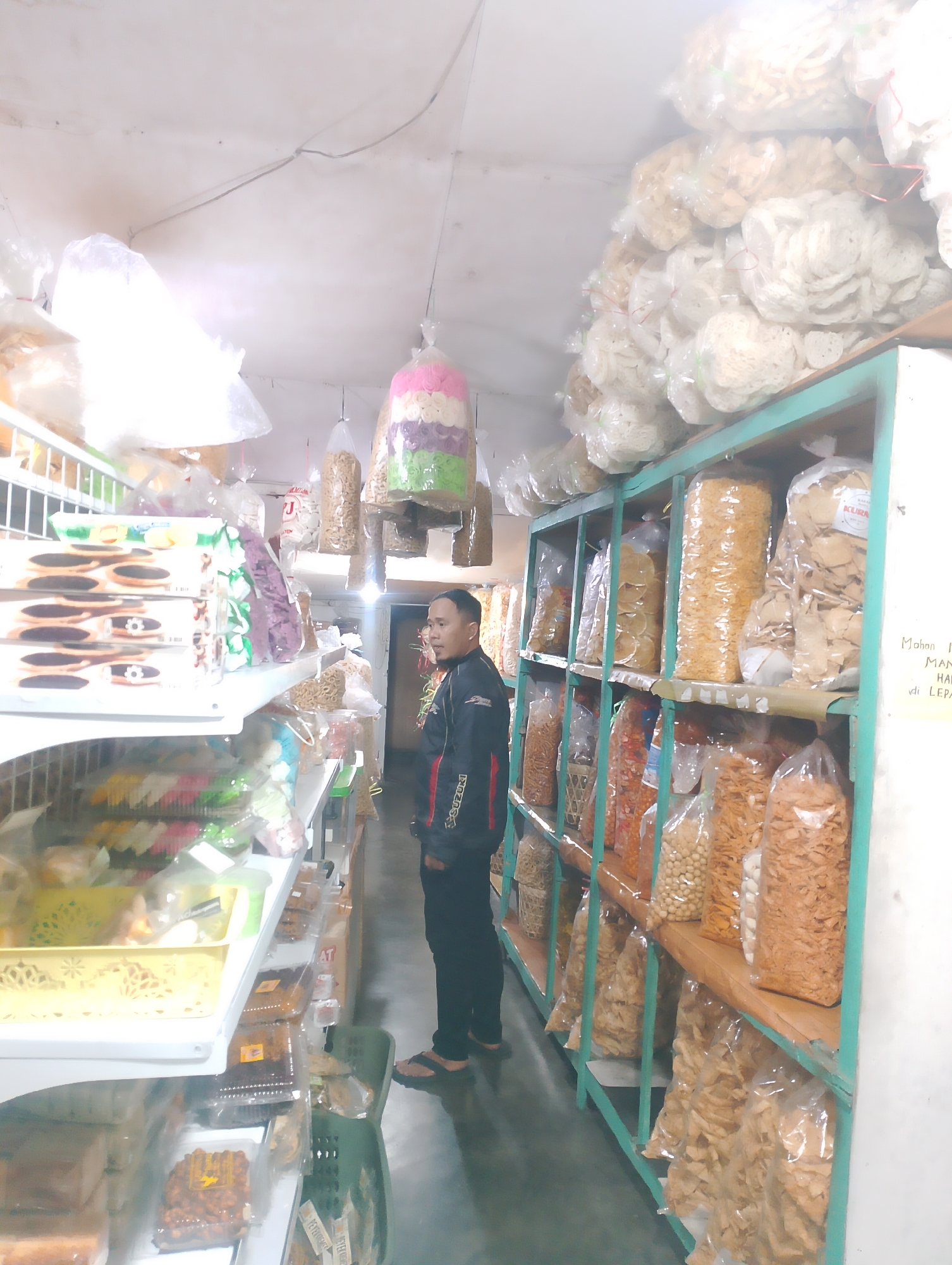In a brightly lit shop in Pakem, Indonesia, a man stands in an aisle surrounded by shelves stacked with various snacks. The shelves are filled with plastic bags of puffed snacks, crackers, and what appear to be candy or baked goods. The man is wearing a dark jacket with a red and white stripe and black pants. He is looking to his left, towards the shelves of snacks. The shop appears to be a small, local establishment, with goods displayed in a colorful and abundant manner. The ceiling is white and shows some wiring, and the walls are also white. The floor is dark.