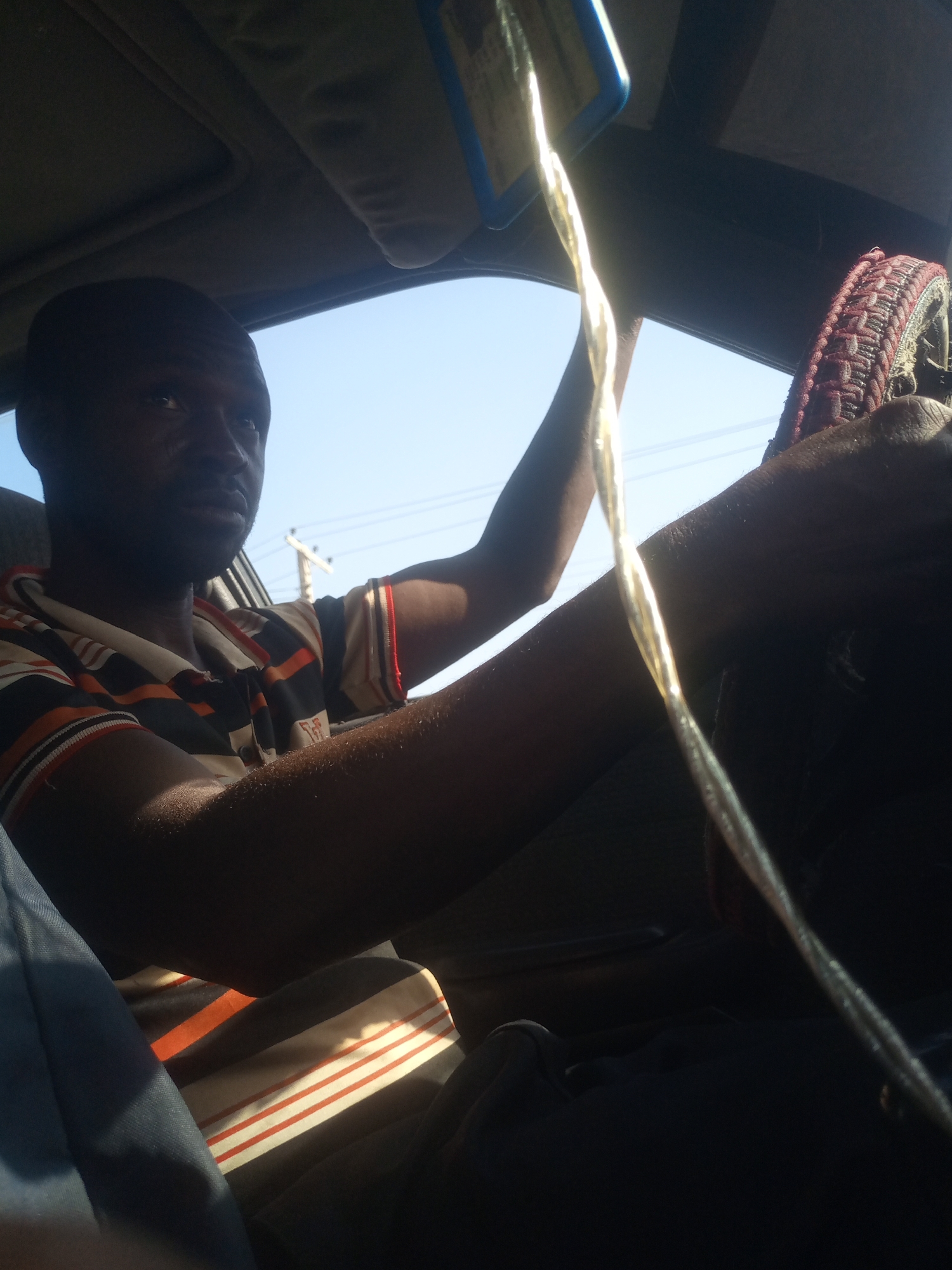 A man is seen from a low-angle perspective inside a vehicle, possibly a taxi, in Gombe, Nigeria. He is behind the steering wheel, which is partially visible with a dark, textured grip and a red and black woven cover. The man, who appears to be of African descent with dark skin, is wearing a horizontally striped t-shirt with orange, white, and black bands. His left arm rests on the steering wheel, and his right arm is extended upwards. A thin, twisted, golden cord or wire hangs from the ceiling of the car, near the rearview mirror, and dangles across the frame, catching the sunlight. The interior of the car is dimly lit, with a bright, sunlit sky visible through the windshield. Power lines and a utility pole are faintly discernible in the background, suggesting an urban or semi-urban setting within Gombe. The overall impression is of a person driving or waiting in a vehicle on a sunny day.