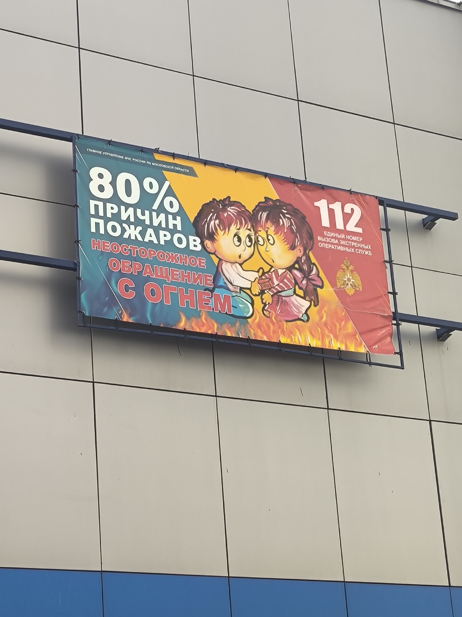 The image shows a sign mounted on a building exterior, likely in an urban area based on the architectural style of the building. The sign appears to be a public safety advertisement related to fire prevention. The text on the sign reads "80% of fires due to careless handling of fire". There are also images of two cartoonish figures surrounded by flames, along with the emergency number 112. The sign also includes text indicating that it is from the Moscow Region branch of the Russian Ministry of Emergency Situations.