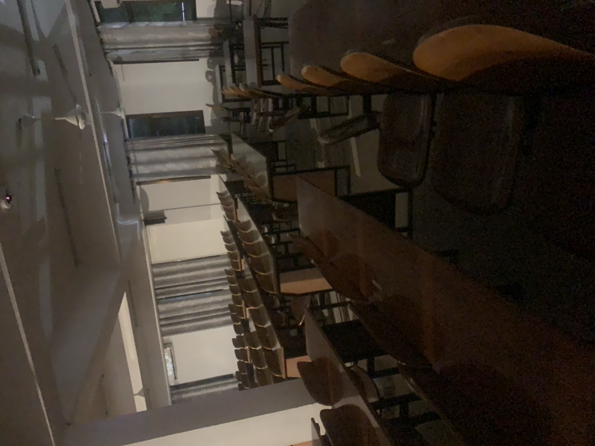 The image shows rows of empty wooden desks and chairs in a tiered lecture hall, likely in a university or educational institution. The room appears to be dimly lit, with light coming in from windows on the left side of the frame. The rows of desks and chairs extend into the distance, creating a sense of depth. The overall impression is of an empty, unused classroom. There is no verifiable information about the city of Dar es-Salaam, Tanzania.