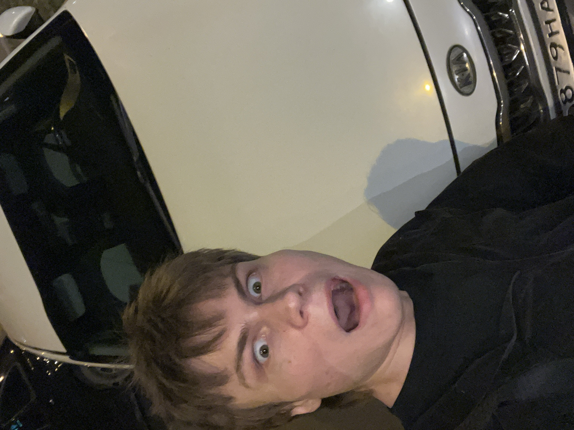 The image is a selfie of a fair-skinned male with brown hair and an open mouth, positioned at an angle relative to the camera. The man is wearing a dark-colored shirt or jacket. A white Kia car is partially visible in the background, with its front end and logo in view. Part of the license plate reads "879HA" which identifies it as a car registered in Moscow, Russia. The image appears to be taken at night or in low-light conditions.