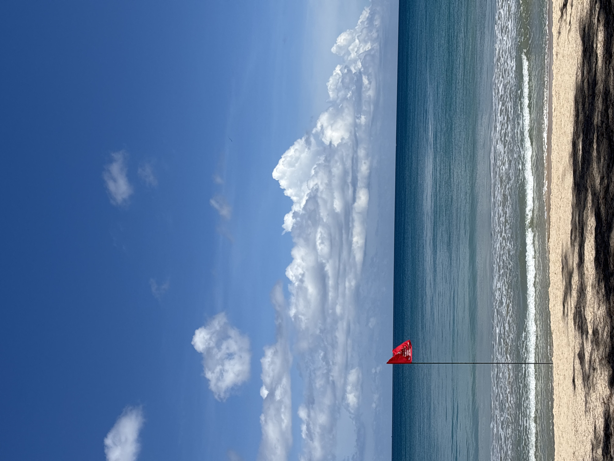The image shows a beach scene, possibly in Choeng Thale, Thailand, judging from the setting. The sky is blue with scattered cumulus clouds. The ocean is a gradient of blue, and the waves are forming near the sandy beach. A red warning flag is planted on the beach. The sand on the beach is light-colored, with dark shadows in the foreground. No people are visible in the image.