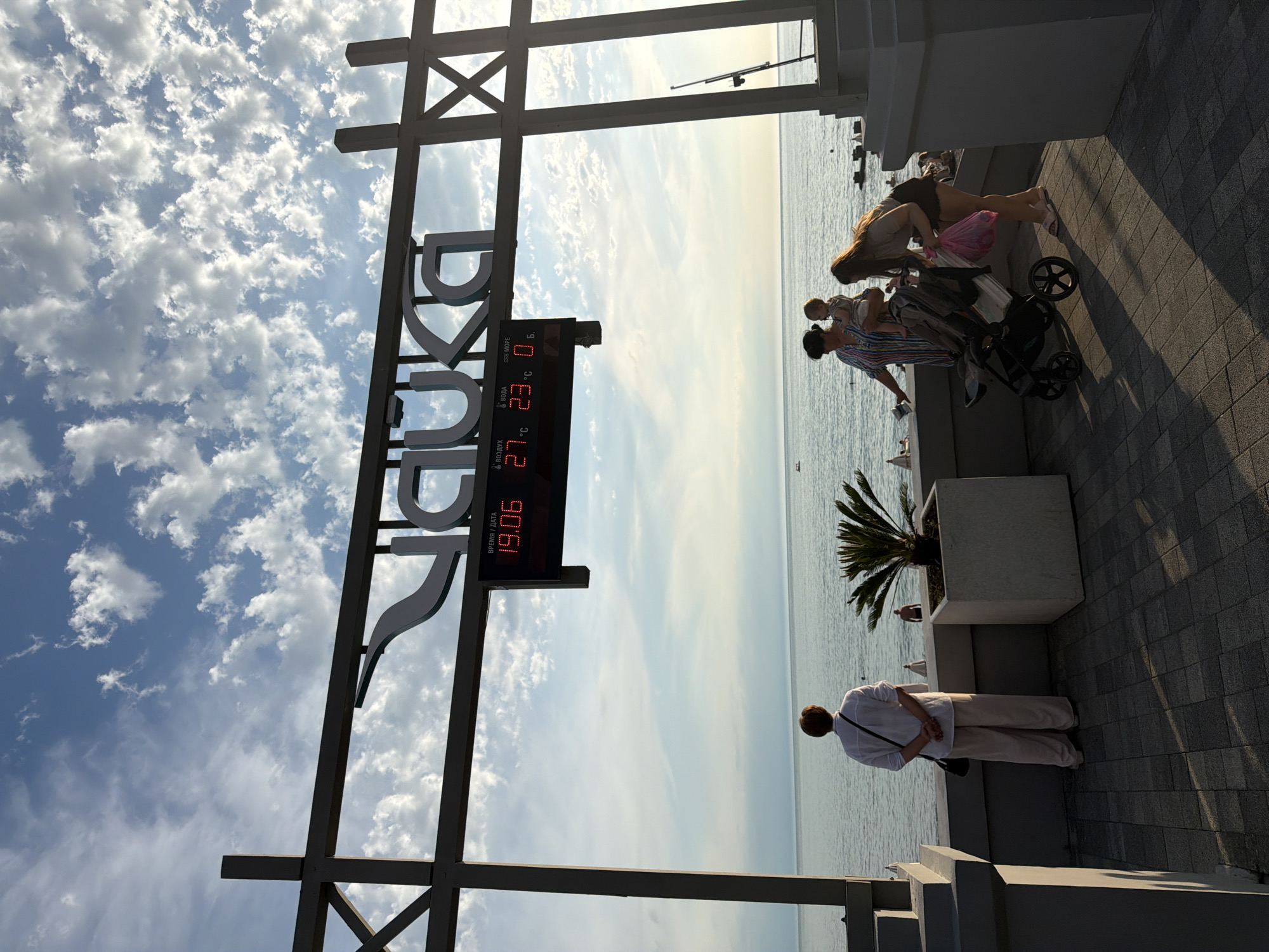 In Sochi, Russia, at the entrance to the "Nauka" science and culture center, a digital display shows the date as June 19th, the air temperature as 27°C, the water temperature as 23°C, and the wind speed as 0 Beaufort. The scene is set on a paved promenade overlooking the sea under a sky filled with scattered clouds.
In the foreground, a woman in a white shirt and light-colored pants stands with her back to the camera, hands clasped behind her. To her right, a planter holds a small palm tree. Further to the right, a group of people are gathered near a stroller. One woman, wearing a striped dress, holds a baby, while another woman in a brown top and skirt bends down, interacting with the stroller and holding a pink bag. A dark-colored stroller is positioned on the promenade.
In the middle ground, the calm sea stretches to the horizon, with a few distant boats visible. The sky above is a pale blue, partially obscured by fluffy white clouds catching the warm light. The overall atmosphere is peaceful and summery, with a few other figures visible on the beach in the distance. The architecture of the "Nauka" entrance, with its prominent sign and metal framework, frames the scene.
