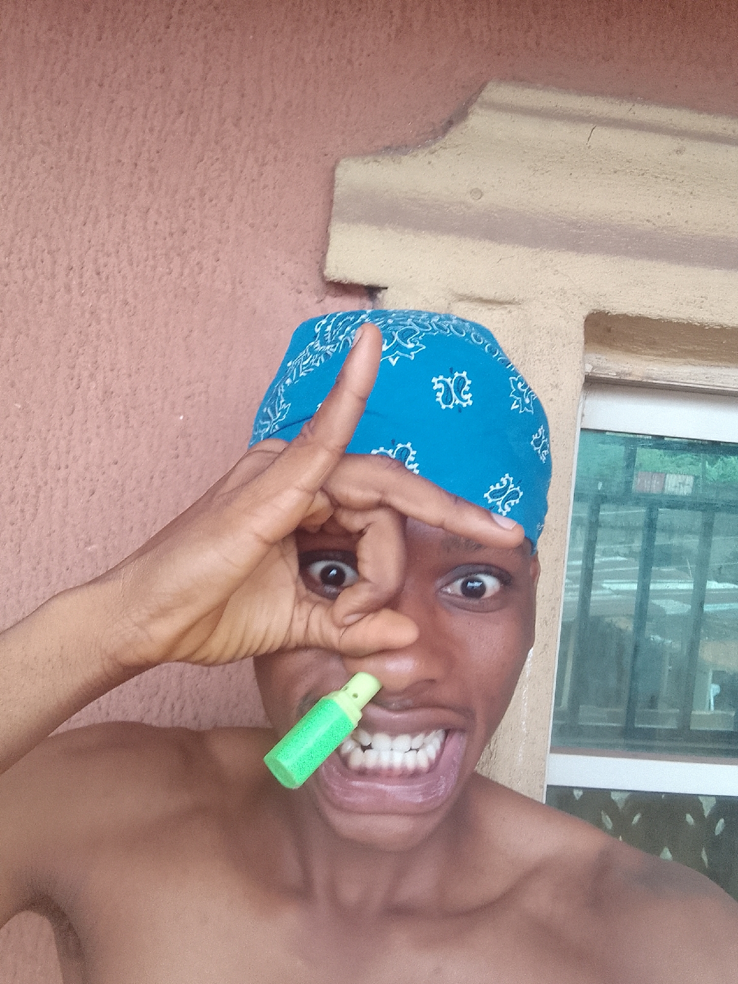A shirtless dark-skinned person wearing a blue patterned bandana is in front of a building with a textured, peach-colored wall and window with vertical bars. The person is making an "OK" hand gesture with their left hand over their left eye. A small, green object is held near their nose. The person is grinning broadly, exposing their teeth. The location is Nkpor, Nigeria.