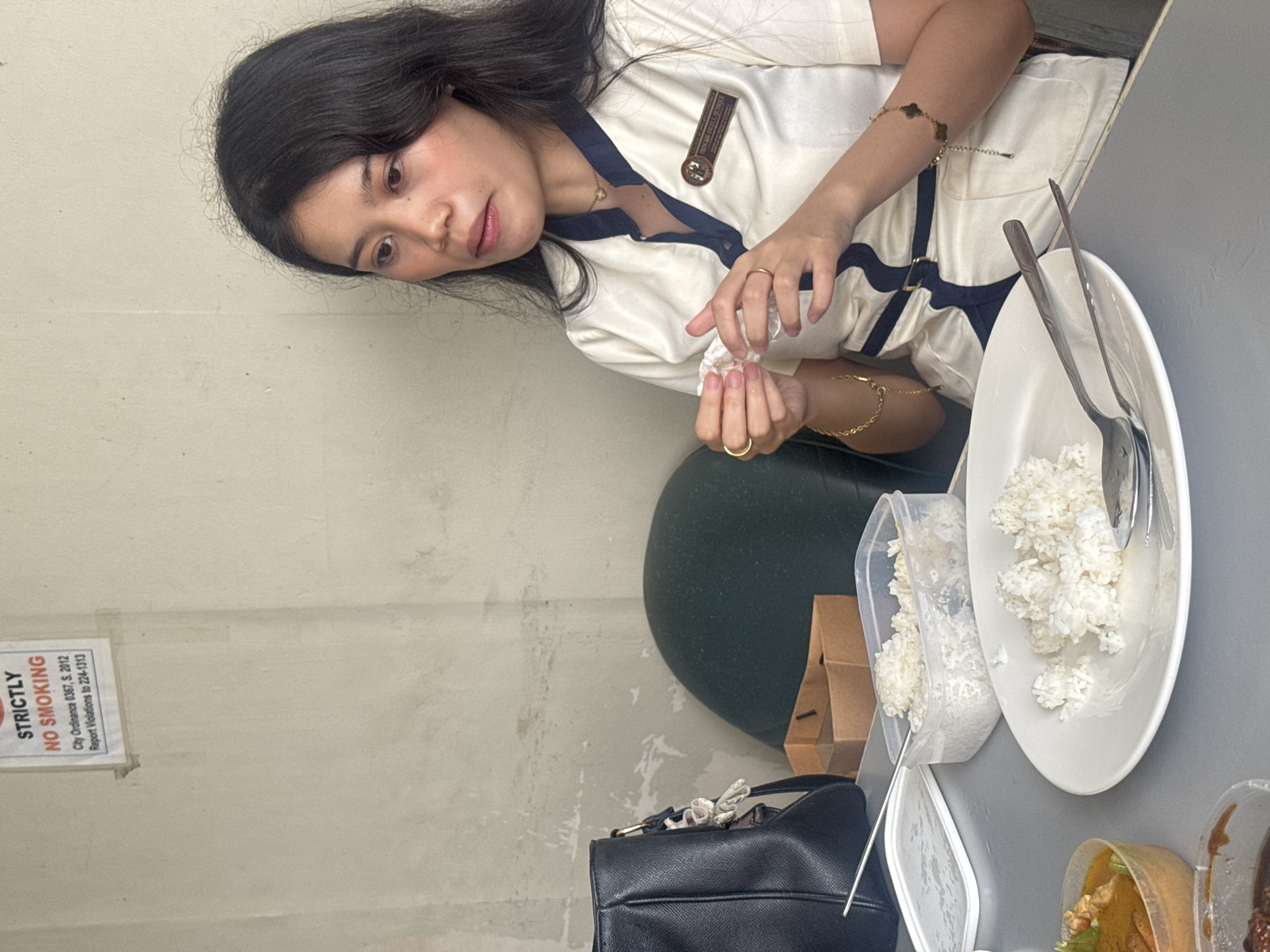 A young woman is seated at a table, holding a white tissue in her hands. She is wearing a white and navy blue uniform with a nametag that reads "PRINCESS ANGELIE QUIAMICO". In front of her are a plate of rice, a plastic container of rice, a brown paper bag, and a black handbag. The wall behind her has a "STRICTLY NO SMOKING" sign, indicating the location is in Davao City, Philippines. The scene appears to be a casual dining or break area.