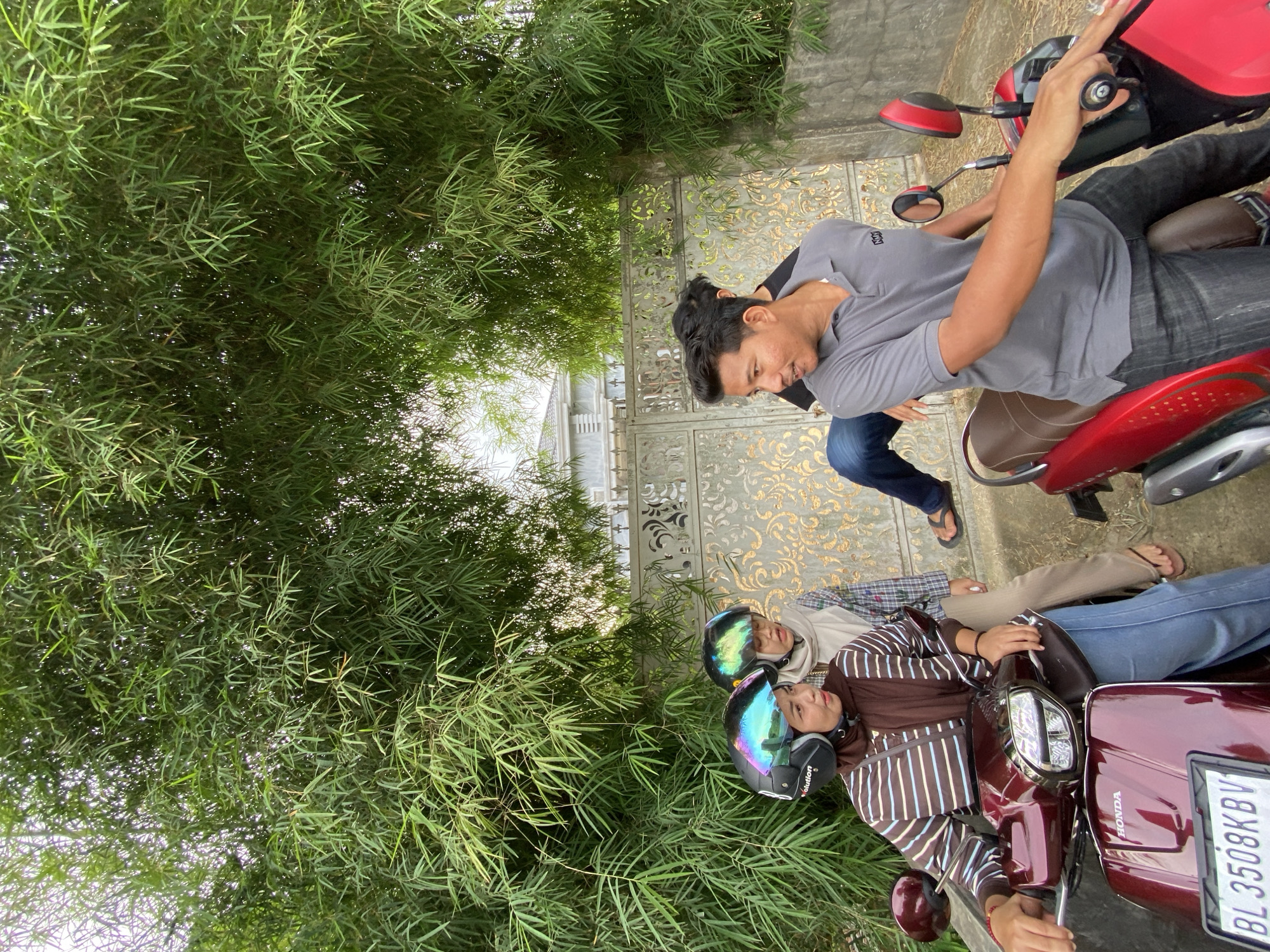 Four people are on two scooters in an outdoor setting, possibly in Meunasahlhok, Indonesia. On the front scooter, a man in a grey t-shirt and dark jeans is riding. Behind him, a person wearing a hijab and a brown and white striped shirt is seated on the scooter. On the second scooter, two individuals are visible, both wearing helmets. One person is in the front, and another is behind them. The scene is surrounded by lush green bamboo foliage, suggesting a tropical or subtropical environment. The scooters are parked on a paved surface, and a decorative metal gate can be seen in the background, hinting at a residential or private property. The lighting suggests it is daytime. The license plate on the rear scooter has visible text: "BL 3508KBV". The overall mood appears casual and relaxed.
