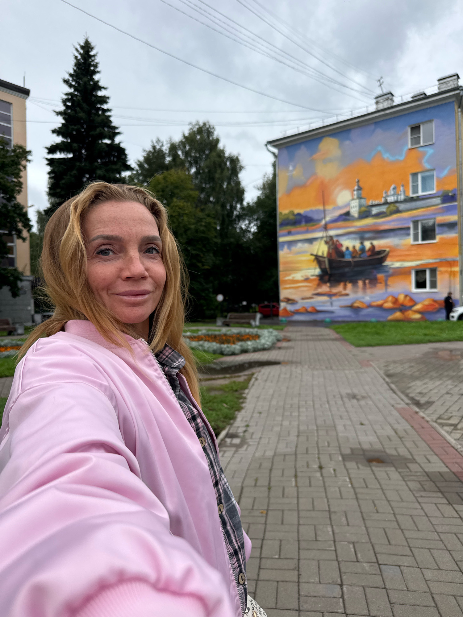 A woman with blonde hair and light blue eyes smiles at the camera, taking a selfie outdoors. She is wearing a pink bomber jacket over a plaid shirt. In the background, a building is adorned with a vibrant mural depicting a sunset over a body of water with a boat and figures. The mural also includes a depiction of a white church with domes. The scene is set in a paved courtyard with a path, flower beds, trees, and a building with windows. This location is identified as being in the city of Veliky Novgorod, Russia.