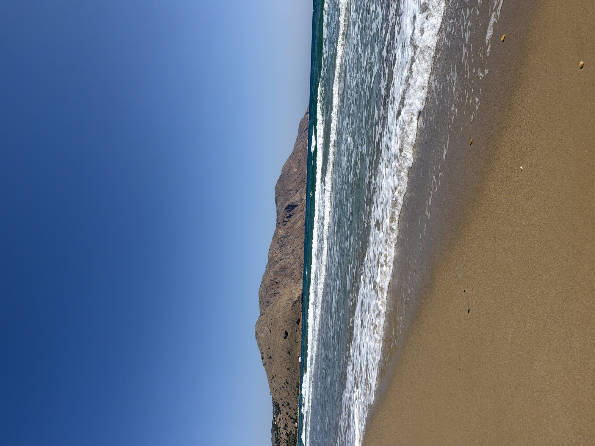 The image depicts a tranquil beach scene on a clear, sunny day in Georgioupoli, Greece. The foreground is dominated by a sandy beach, with gentle waves lapping at the shore. The water is a clear turquoise, transitioning to a deeper blue further out. In the mid-ground, several rows of white-capped waves are visible, indicative of a calm sea. Beyond the water, a large, rugged hill forms the backdrop, its slopes covered in dry vegetation. The sky above is a cloudless, vibrant blue. The overall impression is one of natural beauty and serenity.