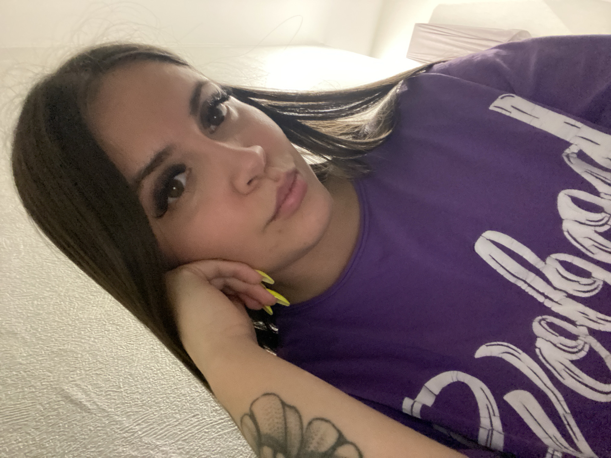 A young woman with dark brown hair and fair skin is lying down, resting her face on her left hand. She has long eyelashes and appears to be wearing makeup. Her fingernails are painted a bright yellow. On her left forearm is a tattoo of what appears to be a flower. She is wearing a purple t-shirt with white writing. The background is a beige wall. A lamp is visible in the upper right corner of the image. There is no information to suggest the location is in the city of Chaikovsky, Russia.