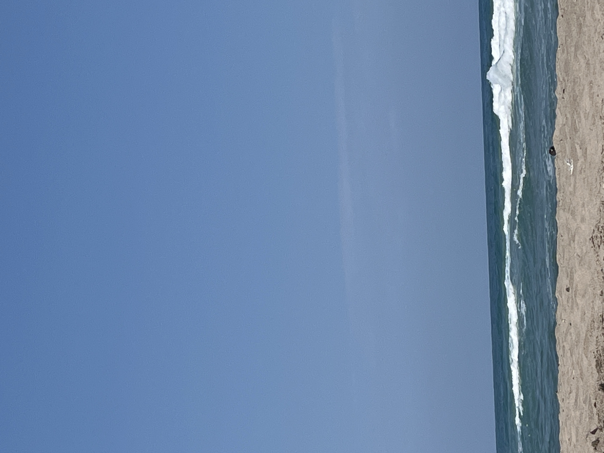 The image depicts a serene beach scene in Ahtopol, Bulgaria. The foreground is dominated by a stretch of sand, showing faint textures and a few scattered small objects, possibly shells or debris. A narrow strip of the sea follows, characterized by its deep blue-green hue and gently rolling waves that break with white foam near the shore. The horizon line is sharp, separating the water from a vast, clear, pale blue sky that occupies the majority of the frame. A subtle, wispy cloud formation is visible in the upper portion of the sky. The overall impression is one of tranquility and natural beauty on a bright day.