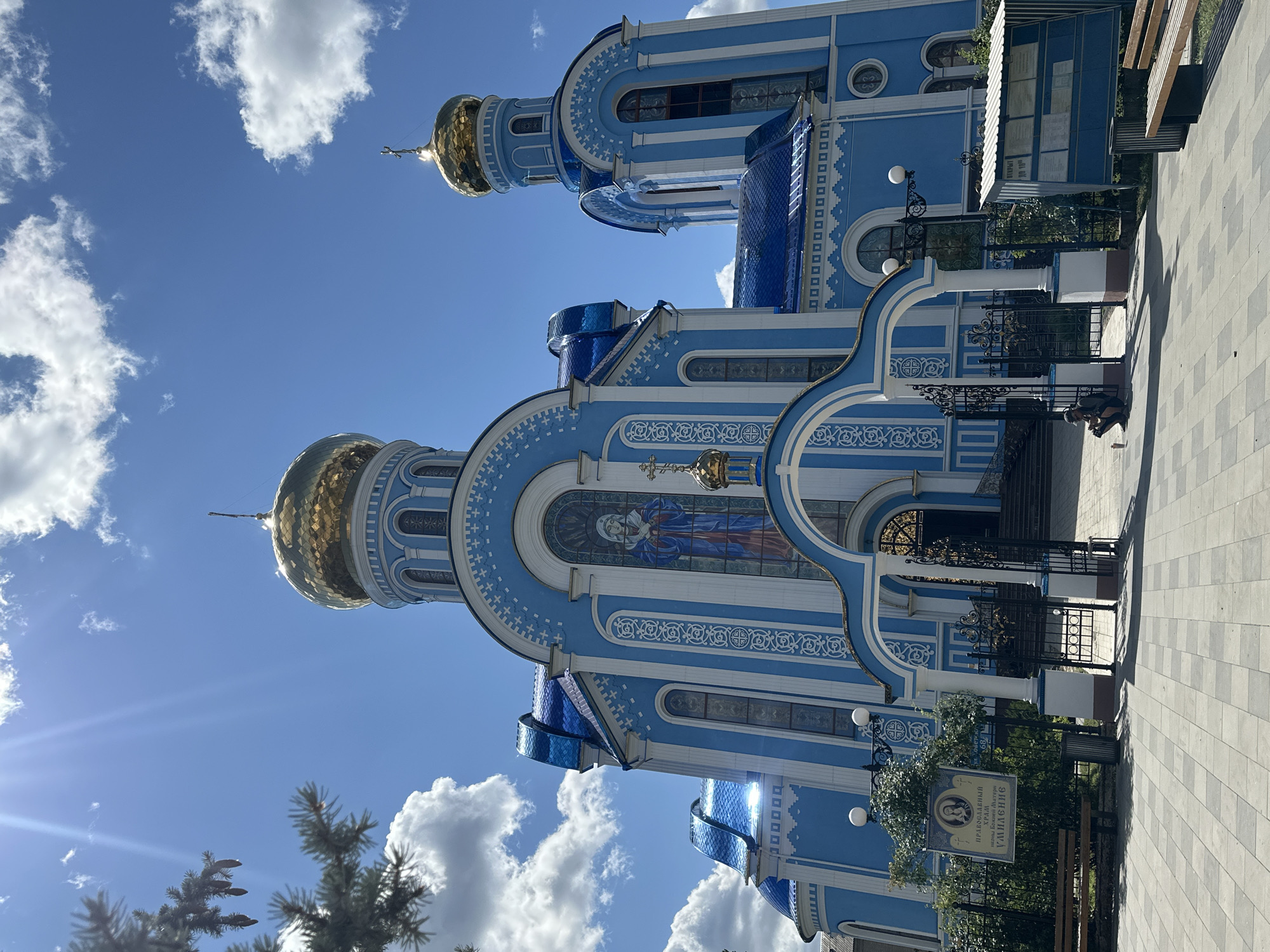 This is a photograph of the Church of the Icon of the Mother of God "Joy of All Who Sorrow" in Raivka, Ukraine, taken on a sunny day. The church is a vibrant blue with white ornamentation and features golden domes. In the foreground, a paved walkway is visible, with benches and a trash can to the right. To the left, a fence encloses a small garden area with a bench and a sign with an icon and Cyrillic text. A single person, wearing a cap and shorts, is kneeling on the ground near the entrance to the church, holding what appears to be a camera or recording device. The sky above is bright blue with fluffy white clouds, and the sun is shining brightly, casting shadows. A tree branch with needles and cones is visible in the upper left corner of the image.