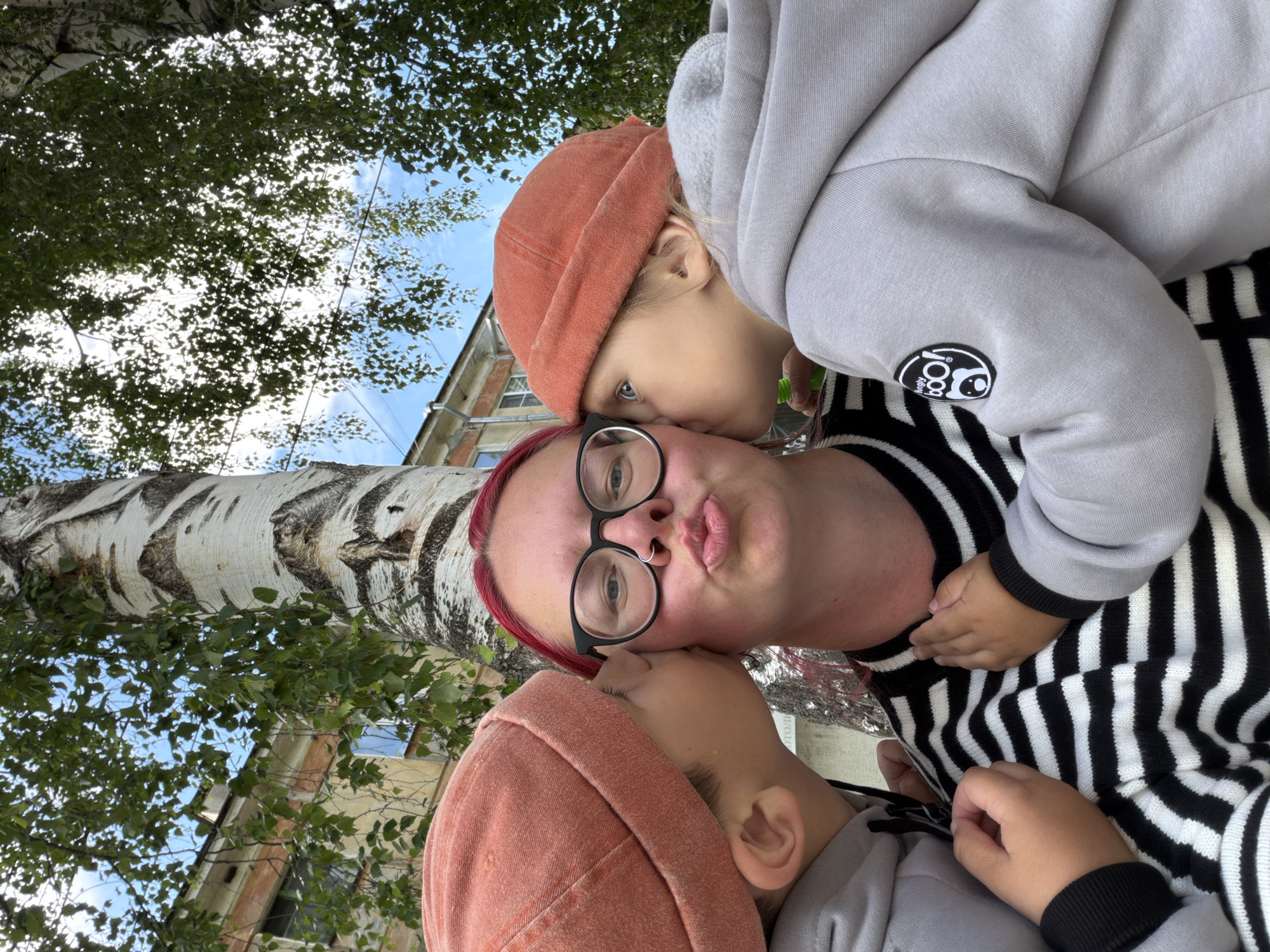 A selfie shows a woman and two children outdoors, with a birch tree and a building in the background. The woman, with red hair and glasses, is leaning her head back, puckering her lips. One child, wearing an orange beanie, is leaning in to kiss her cheek. Another child, wearing a gray hoodie, is also leaning in, with their face partially obscured. The woman is wearing a black and white striped sweater. The children are wearing beanies and hoodies. The sky is blue with some clouds. The leaves of the birch tree are green. The building appears to be residential. The overall emotion is affectionate and playful. The image seems to have been taken during the daytime, likely in the late summer or early autumn, given the foliage. There is no visible text in the image.
