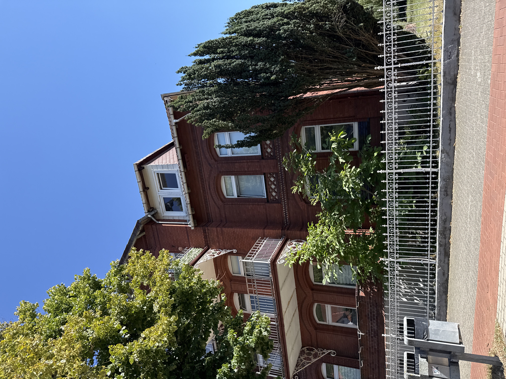A red brick building with white trim and balconies is viewed from a low angle, looking upwards. The building has multiple windows and a sloped roof with a dormer window. In front of the building is a white metal fence. To the left of the building, a large green tree with yellowing leaves is in the foreground, and to the right, a tall, slender evergreen tree stands. The sky is clear and blue. In the lower left corner, there are two gray trash receptacles. The ground in front of the fence is paved with interlocking bricks. This scene is located in Bad Lauterberg im Harz, Germany.