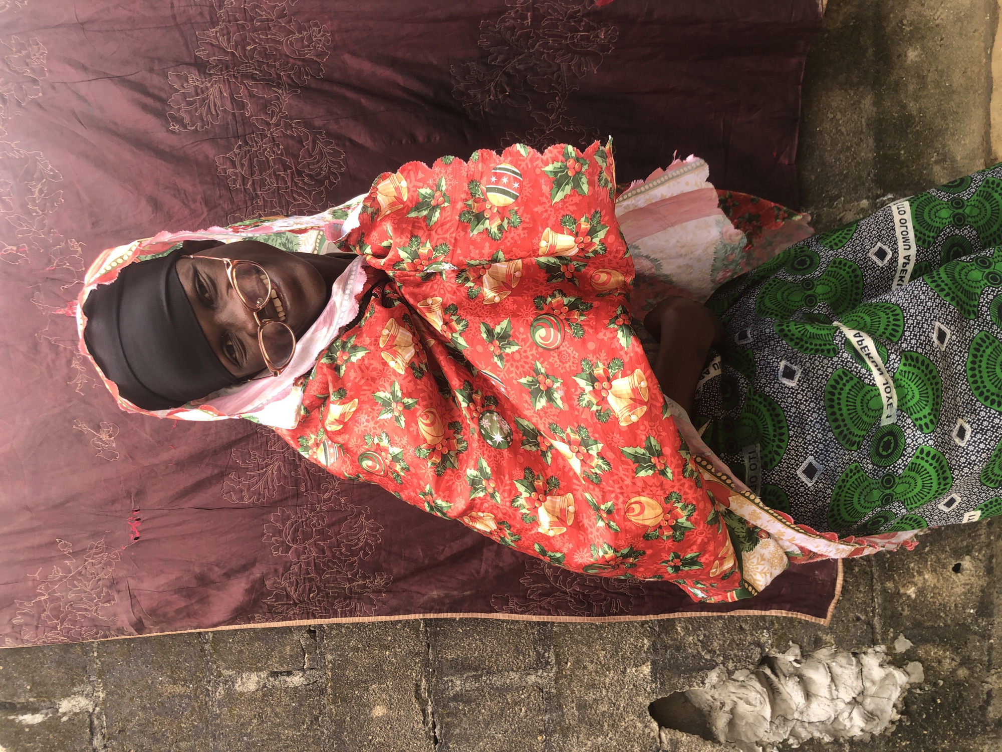 A person wearing a black headscarf, a pink and red patterned fabric draped over their shoulders and a green patterned skirt stands in front of a wall in Ado, Nigeria. The person is wearing rose-gold colored glasses and smiling. The fabric draped over their shoulders has a Christmas-themed pattern with bells, holly, and ornaments. The skirt has a black and green geometric pattern with white diamond shapes. The background consists of a textured, weathered concrete wall and a dark red fabric with floral embroidery.