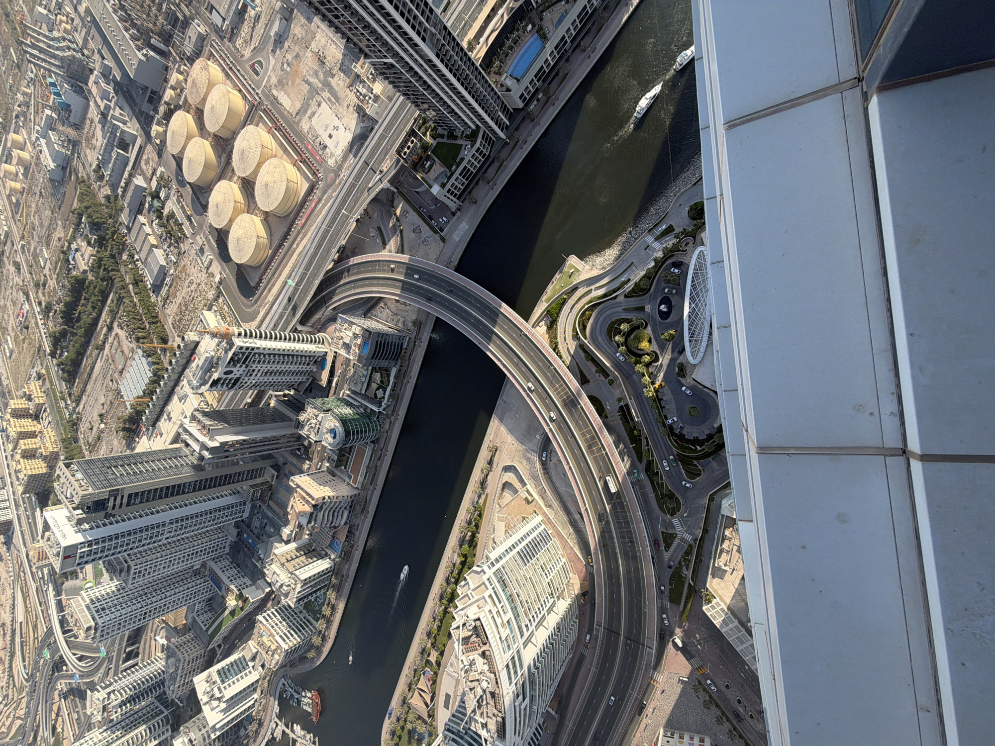 This high-angle, aerial photograph captures a dynamic urban landscape in Dubai, United Arab Emirates, likely taken from a skyscraper looking down steeply. The scene is dominated by a dark, winding canal, flanked by numerous modern high-rise buildings and a complex network of roads, including a prominent curved elevated bridge spanning the waterway.
Several white yachts and smaller boats are visible on the canal, creating subtle wakes, suggesting active water traffic. Cars appear as small specks on the multi-lane roads and the elevated bridge. In one section of the city, on the left, eight large, light-colored cylindrical storage tanks are arranged in a grid pattern within an enclosed area, hinting at an industrial or utility zone. Adjacent to this are other buildings and stretches of undeveloped land, along with some green landscaping and palm trees.
The architecture visible is distinctly modern, characterized by tall buildings with contemporary designs. One building features a unique green-domed structure on its roof. On another building, Arabic text is visible, likely identifying a business or facility. The photograph was taken under bright, clear daylight conditions, indicated by strong sunlight and distinct shadows, suggesting a sunny morning or late afternoon. The right edge of the frame shows part of the light-grey paneled facade of the building from which the image was captured. The overall impression is one of extensive urban development and infrastructure in a bustling modern city.
