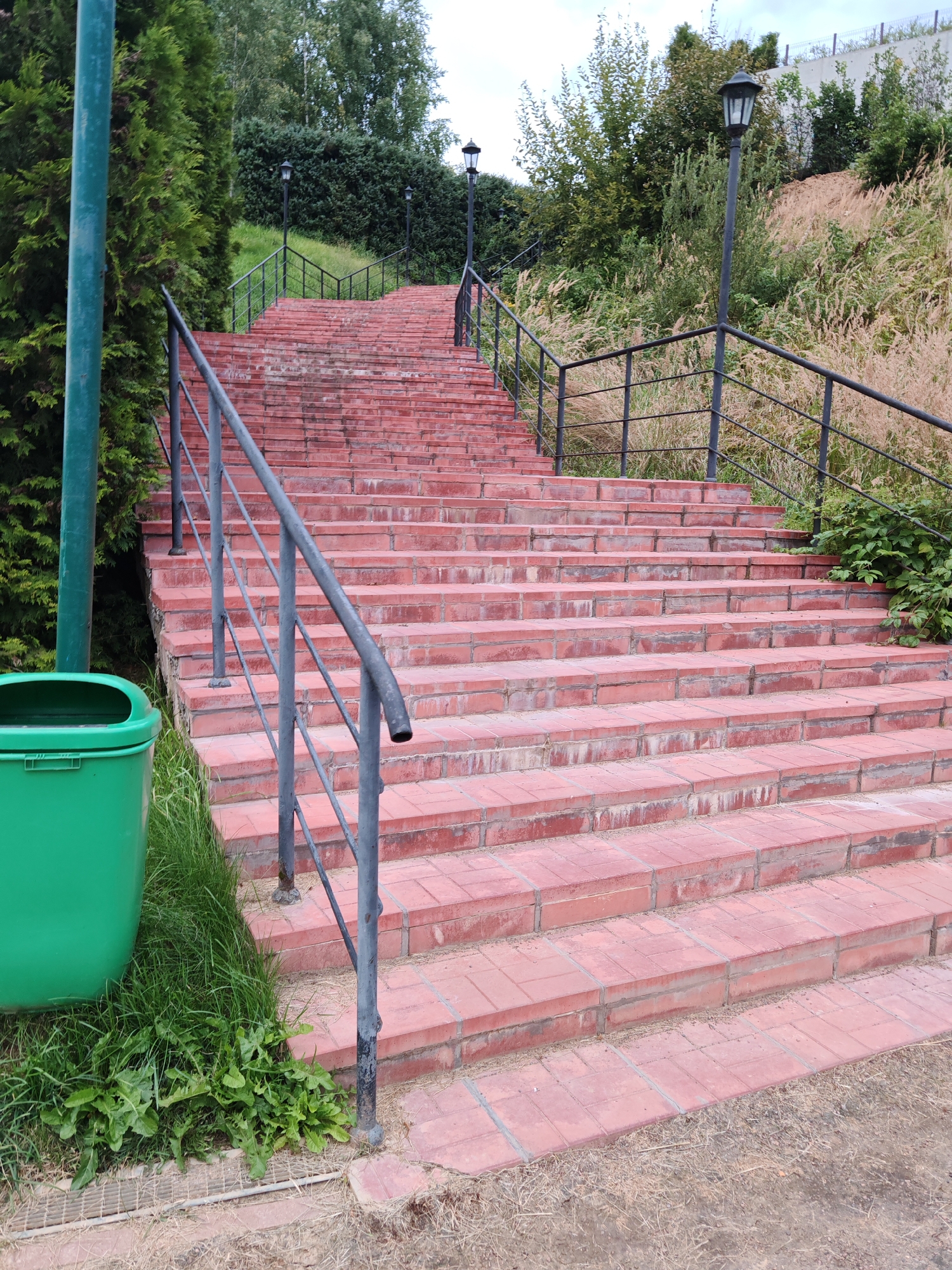 The image shows a long, wide staircase made of red brick, ascending a gentle slope. The stairs are flanked by a dark metal railing on both sides, which also features lampposts at intervals. To the left of the staircase, a tall green metal pole stands next to a bright green trash bin situated among overgrown grass and some weeds. Beyond the railing, the slope is covered in lush greenery, with various trees and bushes, including tall grasses that appear dry and light brown. The sky is overcast. The scene is located in the city of Rosinka, Russia.