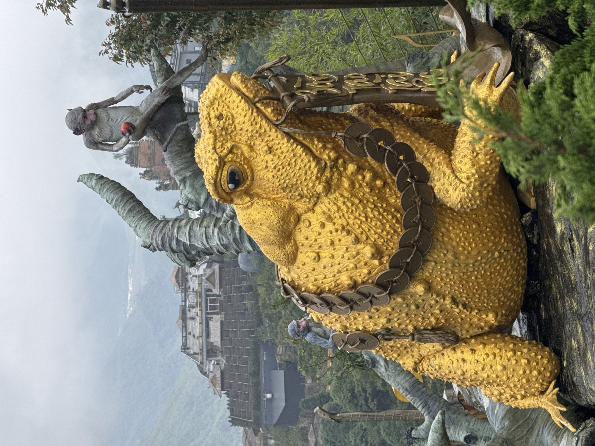 A close-up, medium-shot image reveals a large, golden, three-legged toad statue adorned with a chain of coins around its neck. The statue is located near a monkey statue. The photograph was taken in Binh Lu, Vietnam. The background shows buildings and greenery, suggesting the statue is situated in a mountainous area. The sky appears overcast or foggy, lending a soft diffused light to the scene.