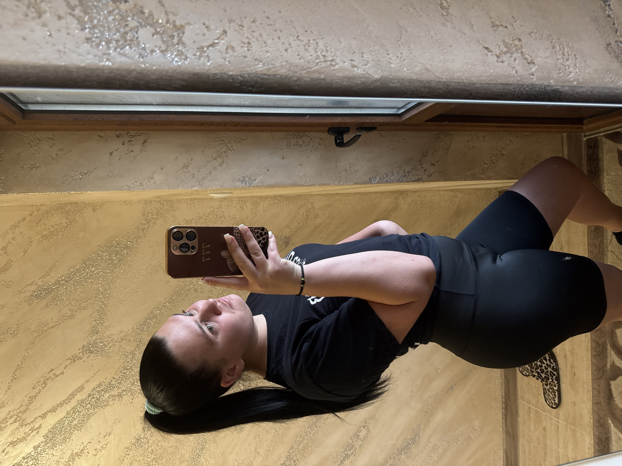 A full-body, mirror selfie shows a woman with dark hair pulled into a high ponytail, wearing a black t-shirt and black shorts. She is holding a brown phone case with a leopard print design. The background is a textured wall with a golden hue. A wooden-framed door is visible to the right. A leopard-print slipper is on the floor to the left. The setting appears to be indoors. There is no indication of the location being the city of Rogosh, Bulgaria.