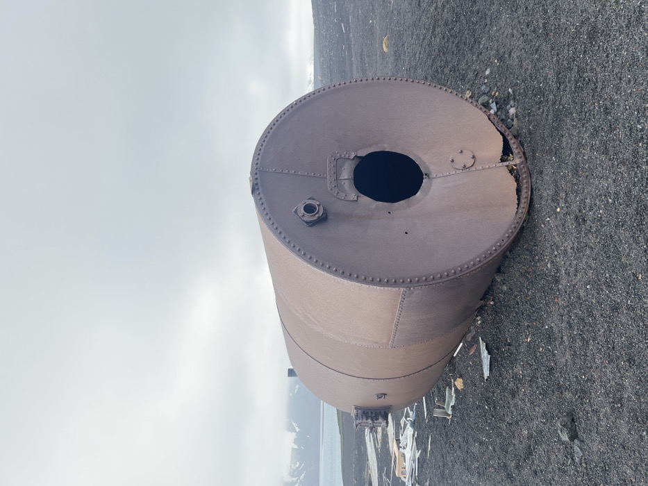 A rusty, cylindrical tank lies on dark, gravelly ground under a cloudy sky. The tank has a large, round hole at one end, with some damage to the surrounding metal. Rivets and a smaller port are visible on the tank's surface. In the distance, a sliver of water and snow-covered mountains are faintly visible. Debris is scattered around the base of the tank.