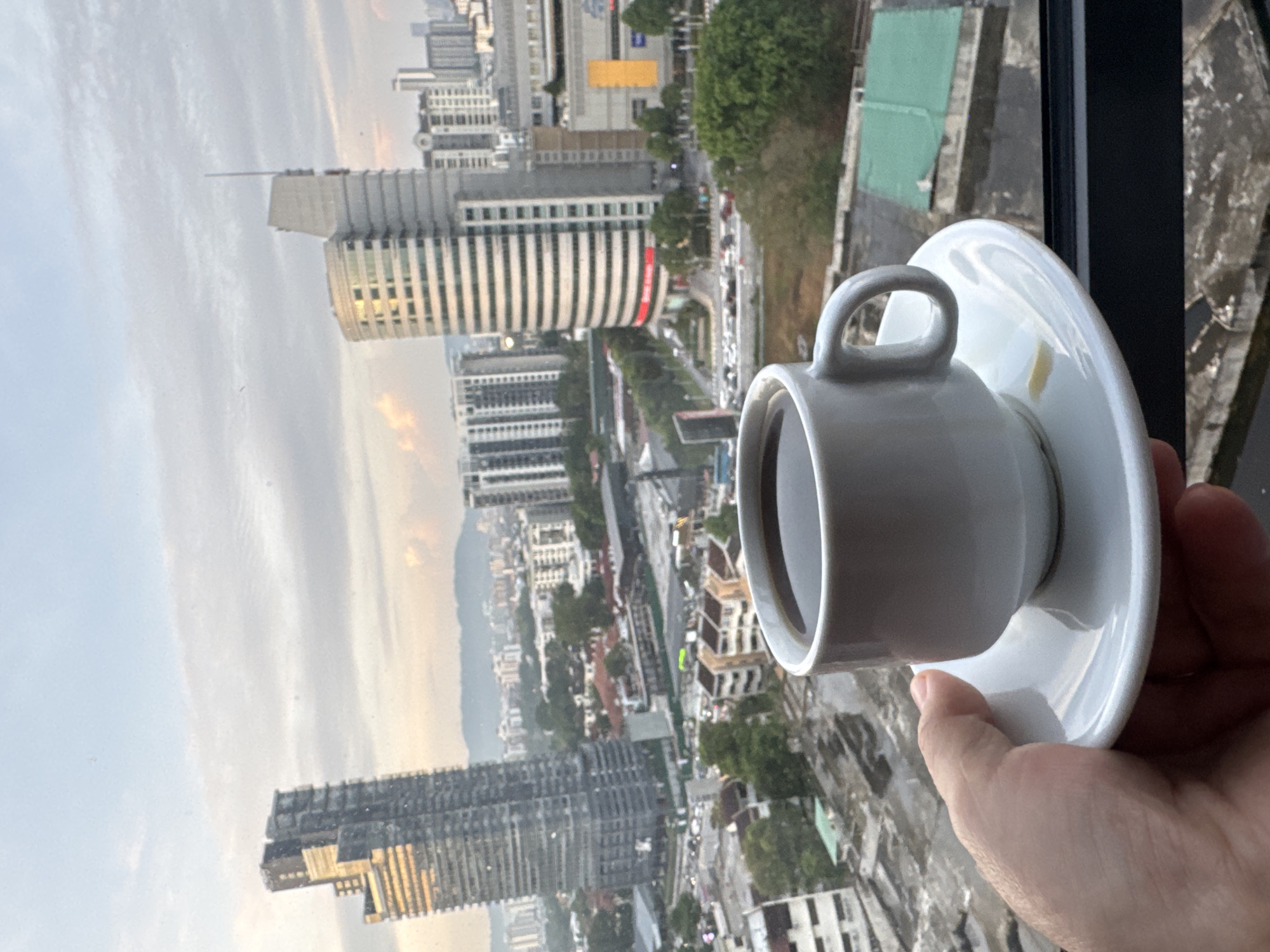 A person's hand holds a white coffee cup and saucer in the foreground, with a panoramic view of the Kuala Lumpur cityscape in the background. The coffee cup is filled with dark coffee. The background features a dense collection of urban buildings, including several high-rise skyscrapers and lower-rise structures, suggesting a bustling city environment. Some greenery is visible between the buildings. The sky above is partly cloudy, with the light suggesting it could be either early morning or late afternoon. The vantage point appears to be from a high floor in a building, possibly a hotel or apartment. The image captures a moment of relaxation with a coffee while observing the urban landscape of Kuala Lumpur.